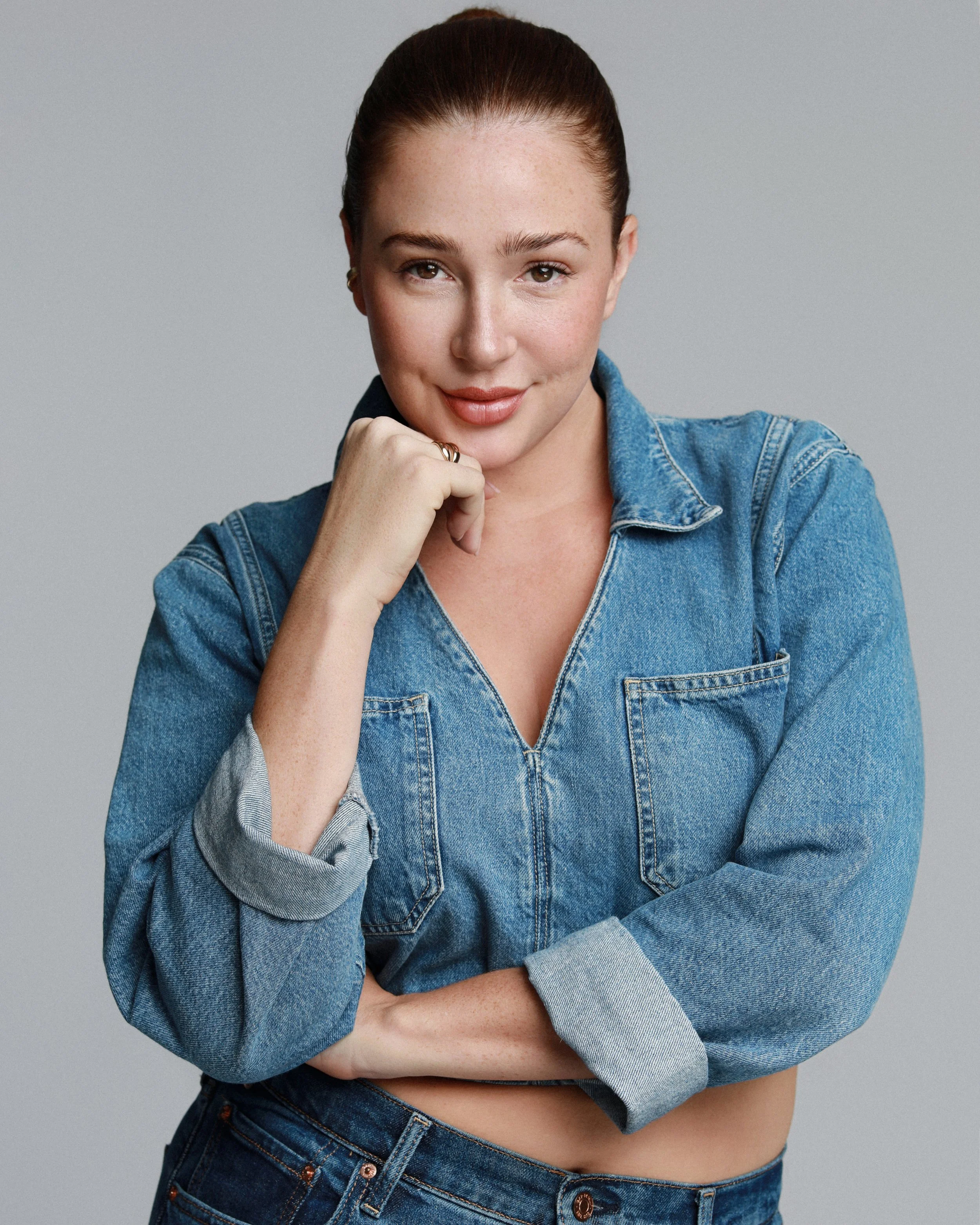 A woman with red hair tied back, wearing a denim jacket and jeans, standing against a plain gray background, with her right hand resting on her chin and a slight smile.