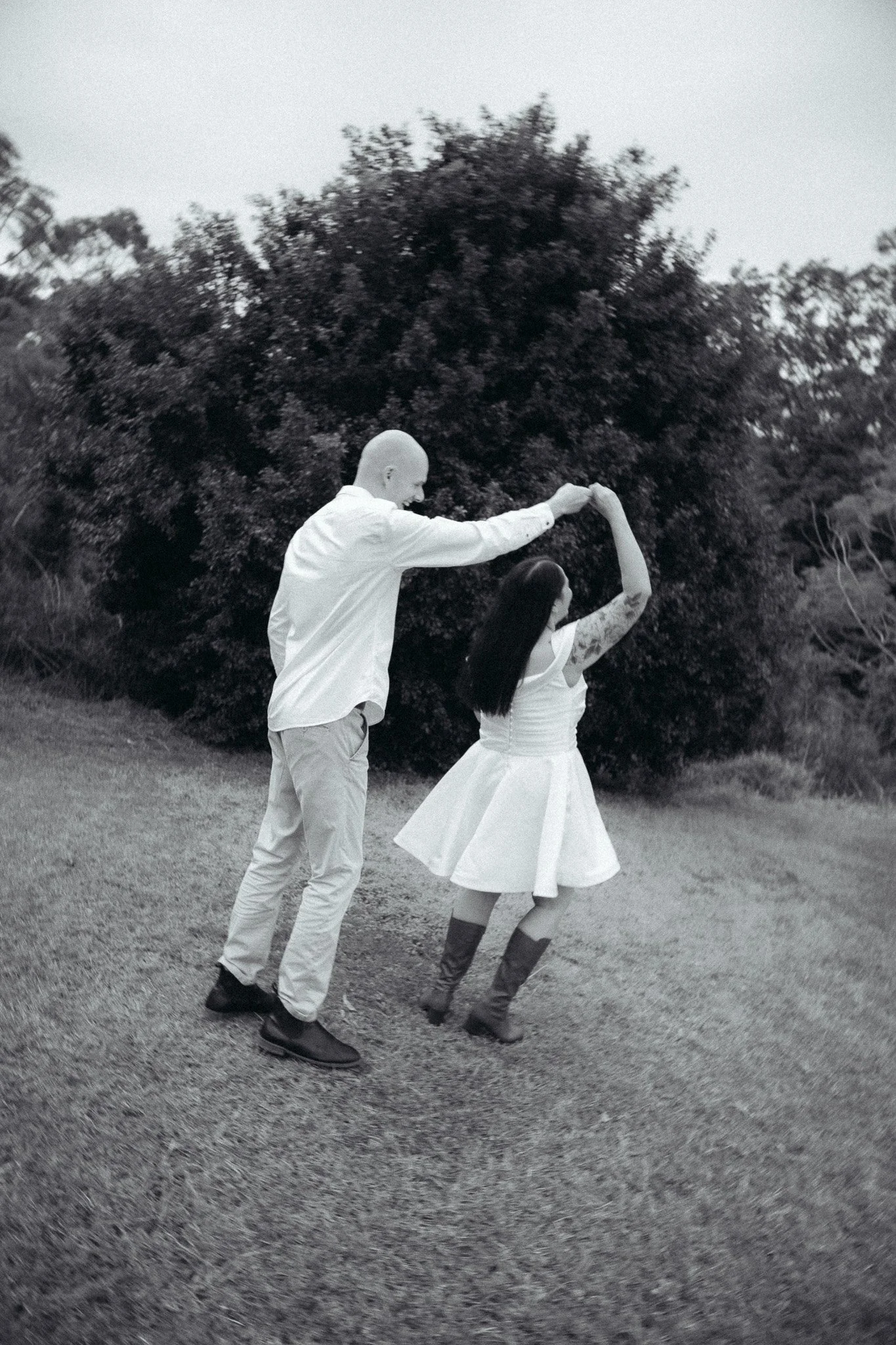 A bald man and a woman with long dark hair dancing outdoors on grass with trees in the background, holding hands as they dance.