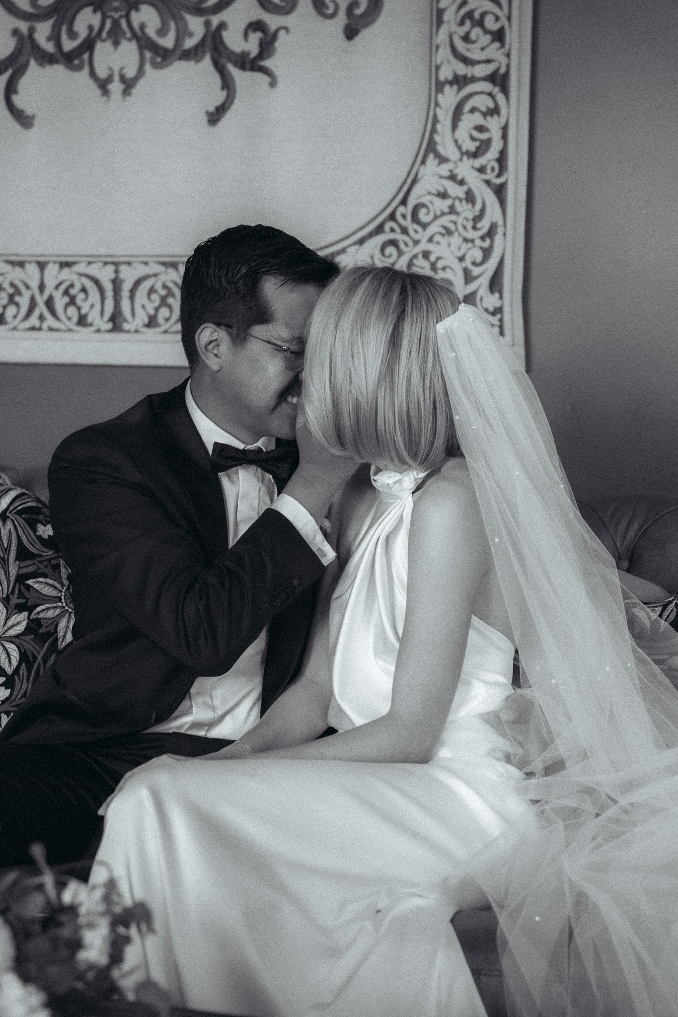 A black and white photo of a bride and groom sitting close, with their foreheads touching. The groom is wearing a tuxedo with a bow tie, glasses, and has dark, styled hair. The bride has short, blonde hair and is wearing a wedding dress with a veil. They are in a decorated room with an ornate wall hanging behind them.