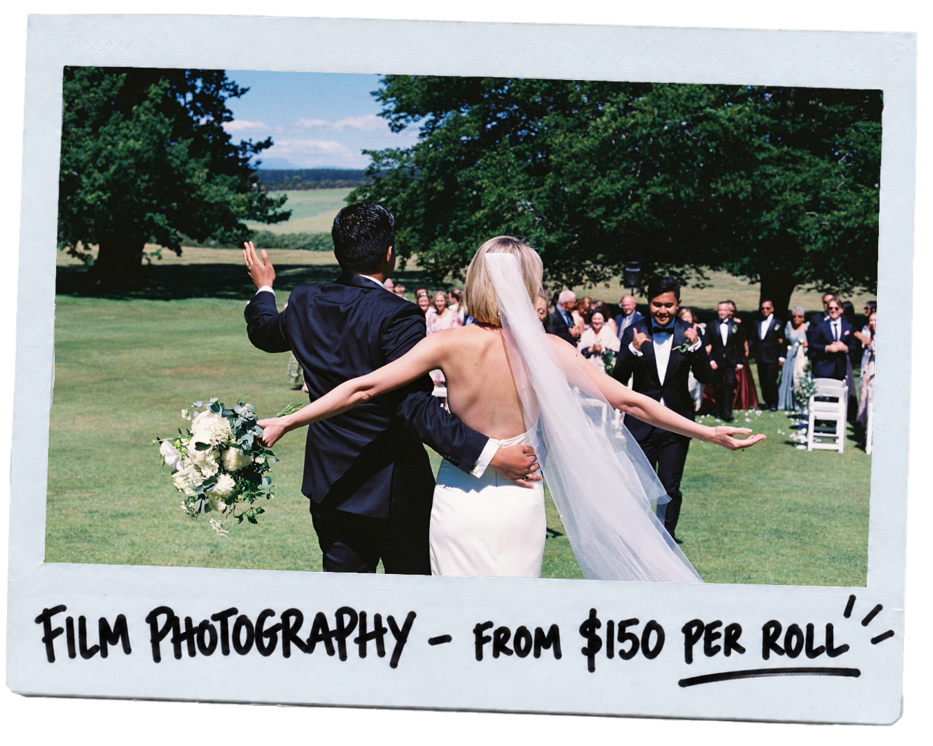 A wedding scene with a bride and groom dancing outdoors on a grassy field, surrounded by wedding guests, with trees and blue sky in the background.