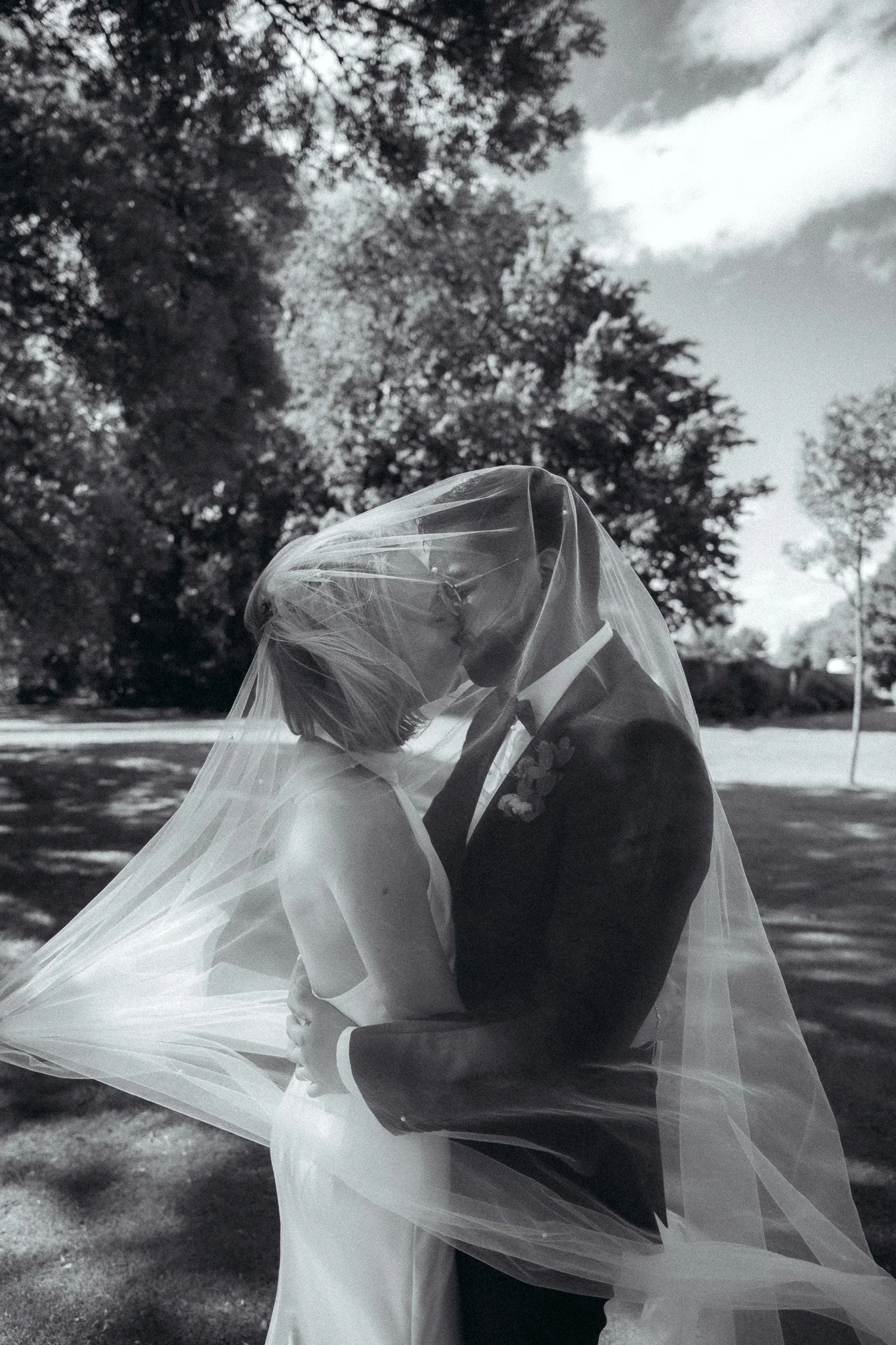 A bride and groom are kissing outdoors, with the bride wearing a veil and a wedding dress, and the groom in a dark suit. Trees and a partly cloudy sky are in the background.