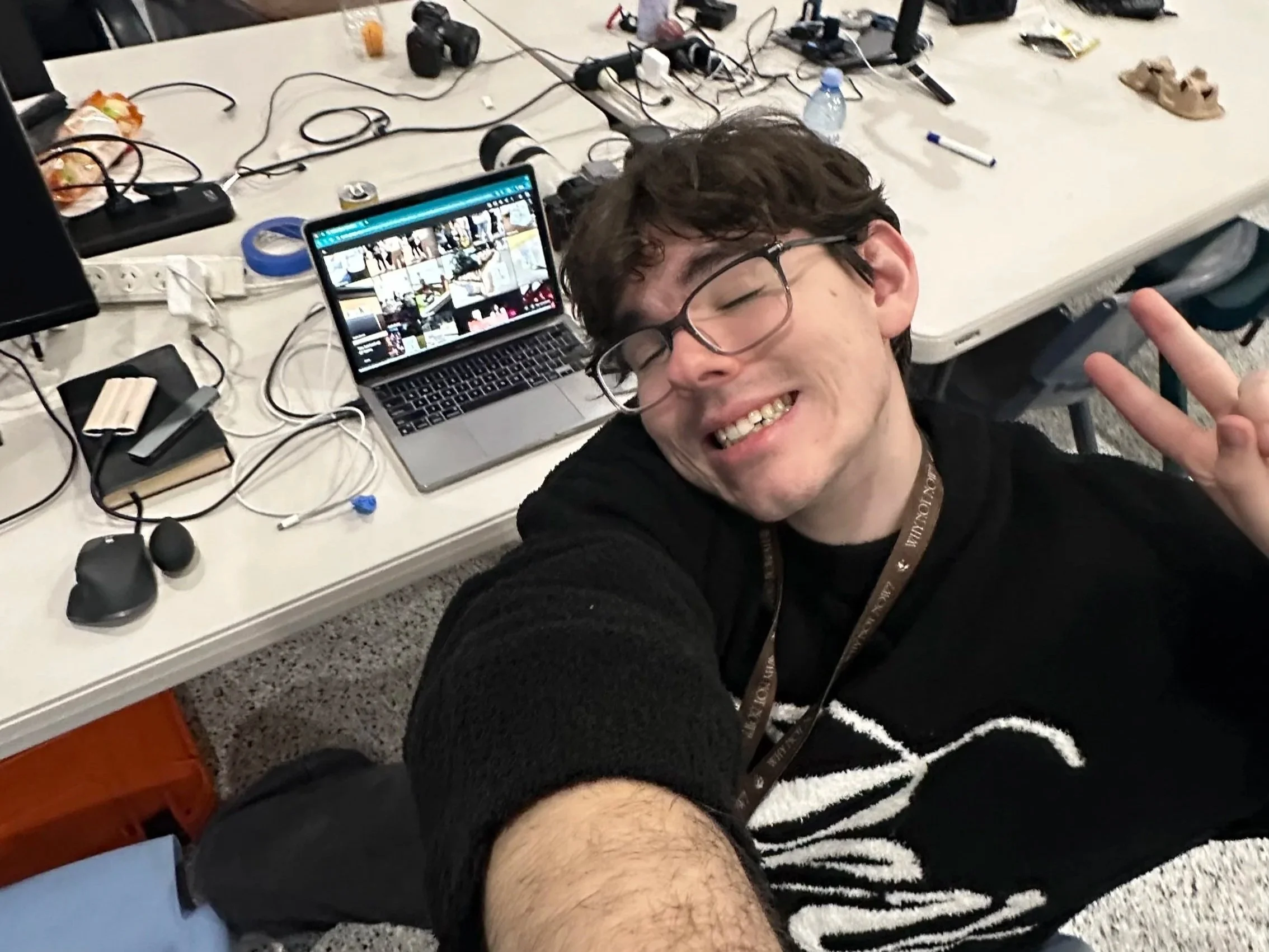 Founder of Joystudios  wearing glasses smiles and makes a peace sign beside a laptop on a cluttered worktable.