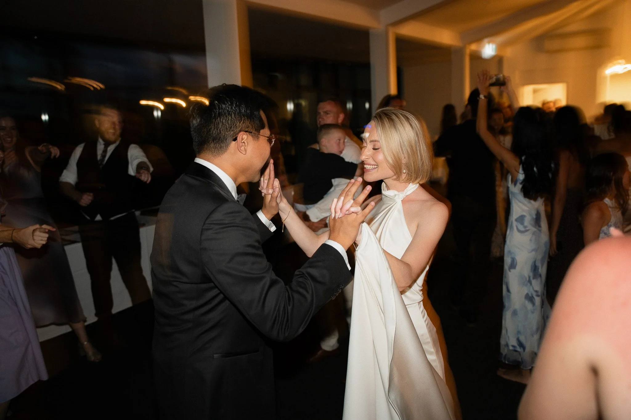 A couple dancing closely at a lively social event with many people in the background, some taking photos and enjoying the moment.