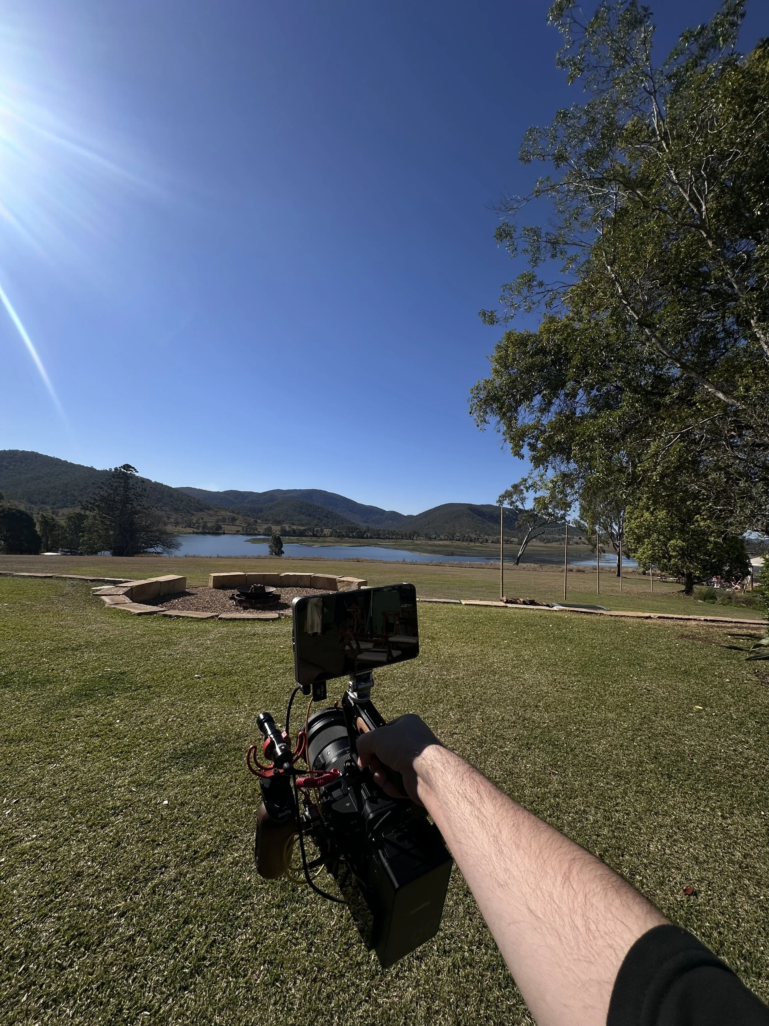 Camera operator filming a scenic mountain lake surrounded by grassy grounds and trees under a clear blue sky