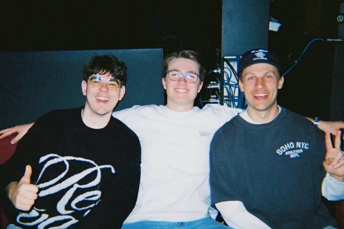 Three smiling men posing together in a recording studio, with audio equipment and cables in the background.