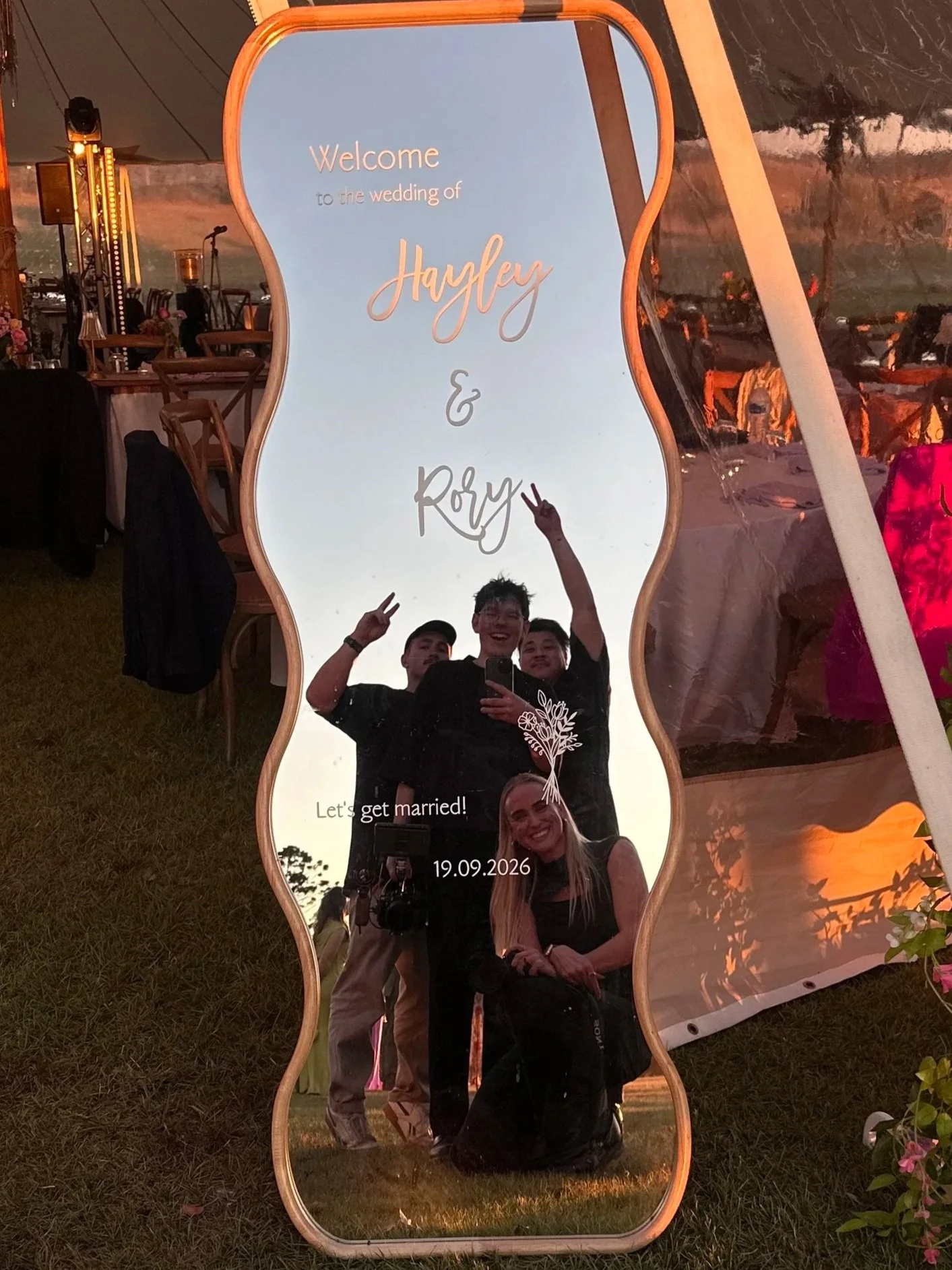 Wedding photo booth mirror reflecting four wedding vendors posing beneath a sign welcoming guests to Hayley and Rory's wedding.