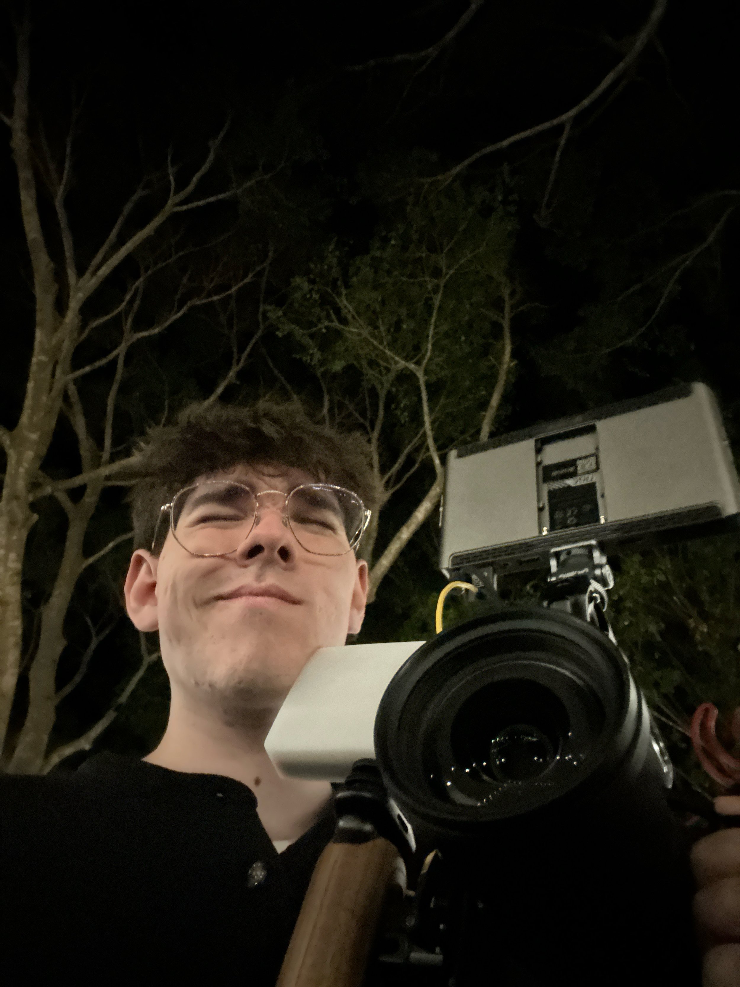 Founder of Joystudios, wearing glasses, holds a large camera with a telephoto lens outdoors at night, with trees and a building behind them.