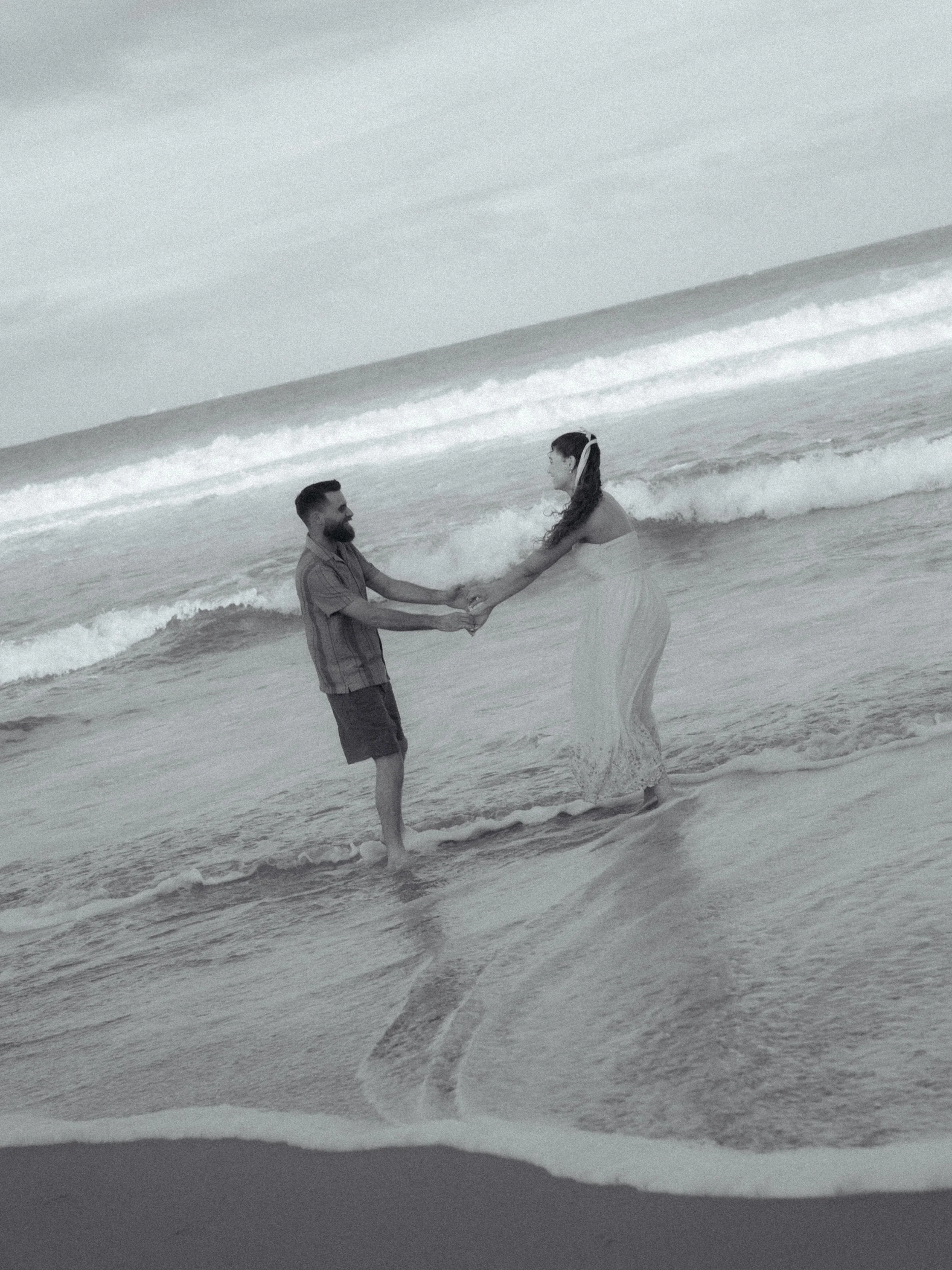 A man and woman are holding hands in the ocean, standing in the water as waves crash around them, during what appears to be a wedding or special event.