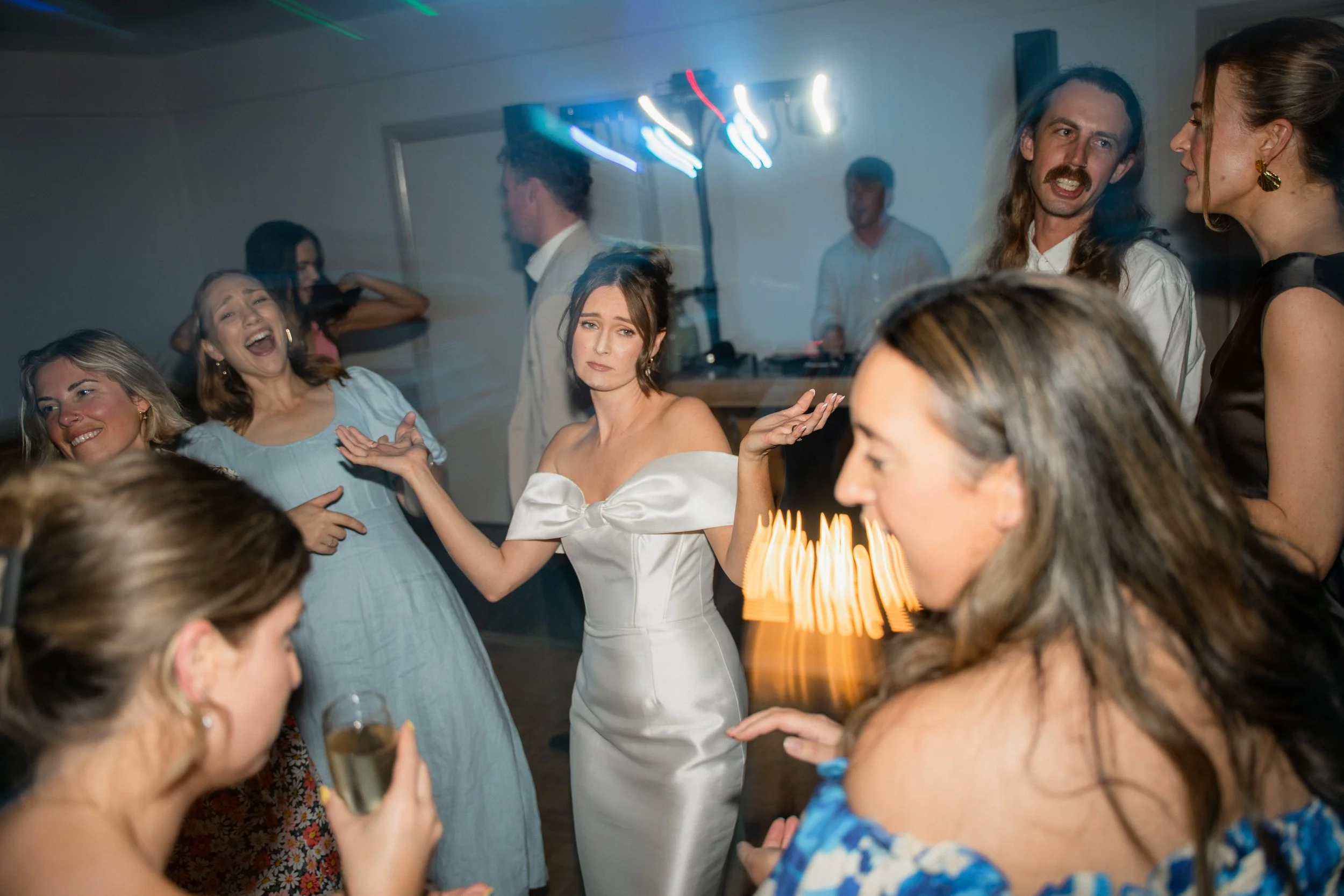 A group of people at a celebration, including a woman in a white dress with a bow, several women are holding drinks, some are smiling, and others are engaged in conversations. The scene appears lively with some motion blur from dancing or movement.