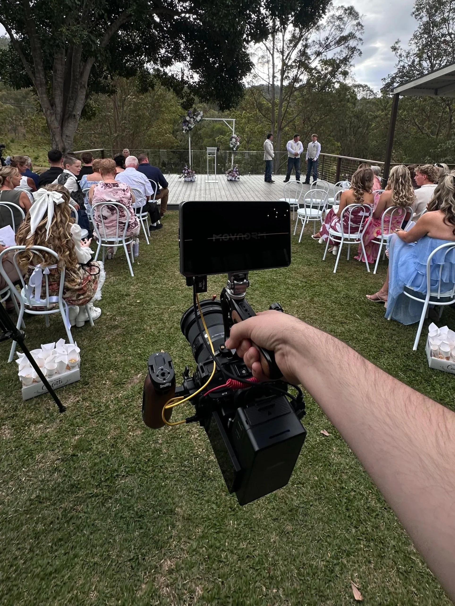 Outdoor wedding ceremony with seated guests, floral altar, and videographer filming in a wooded setting