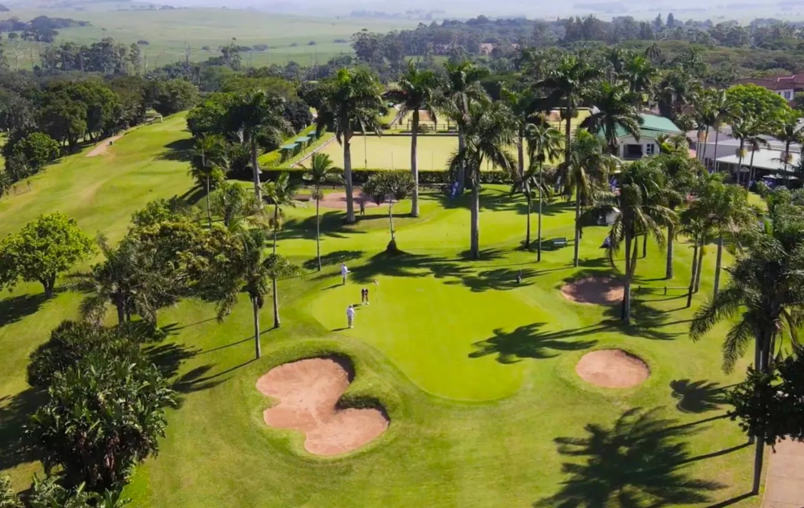 Aerial view of a golf course with green fairways, sand bunkers, palm trees, and players. Surrounding residential buildings can be seen in the background.