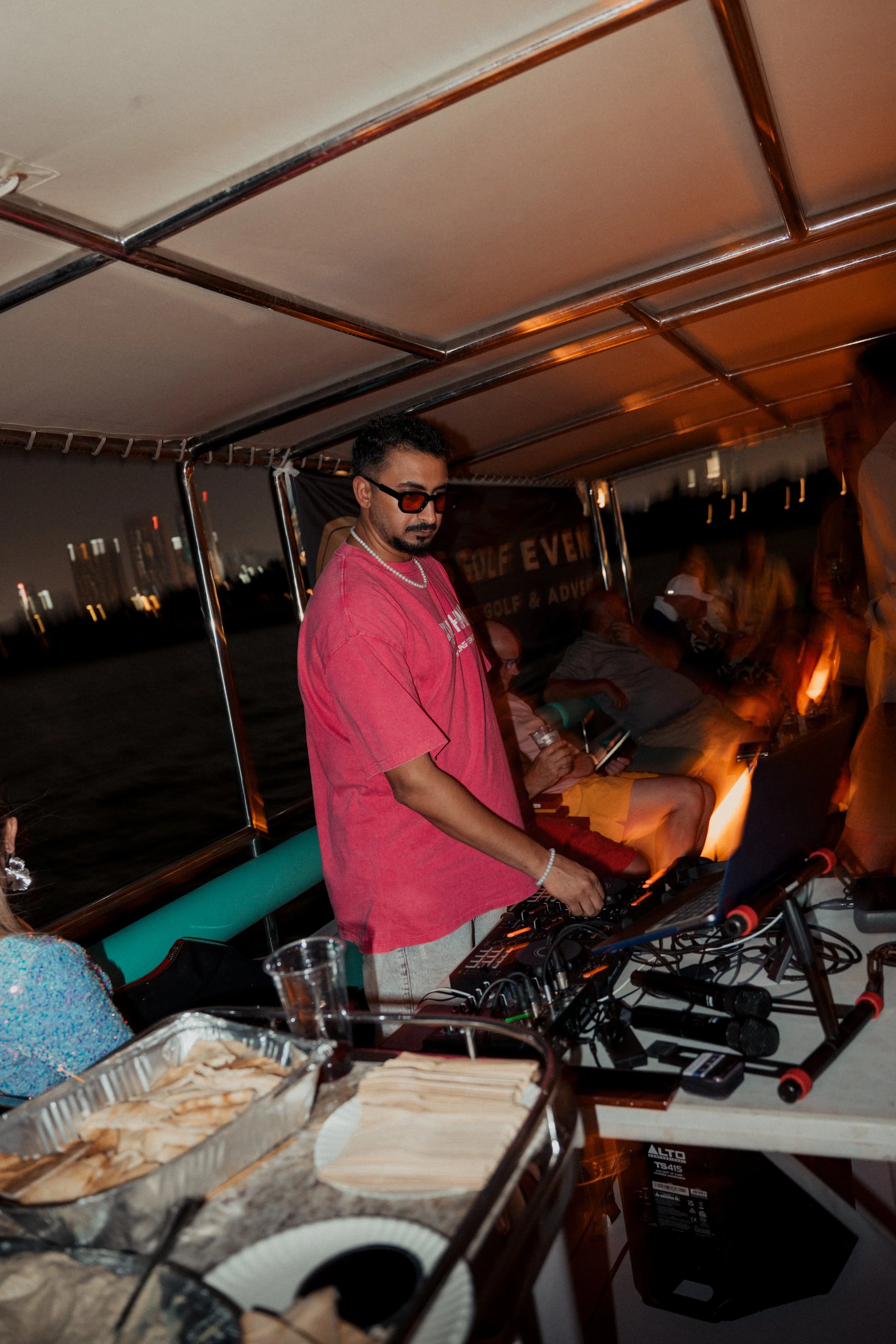Man DJ wearing sunglasses and a red shirt playing music on a boat at night with other people relaxing in the background.
