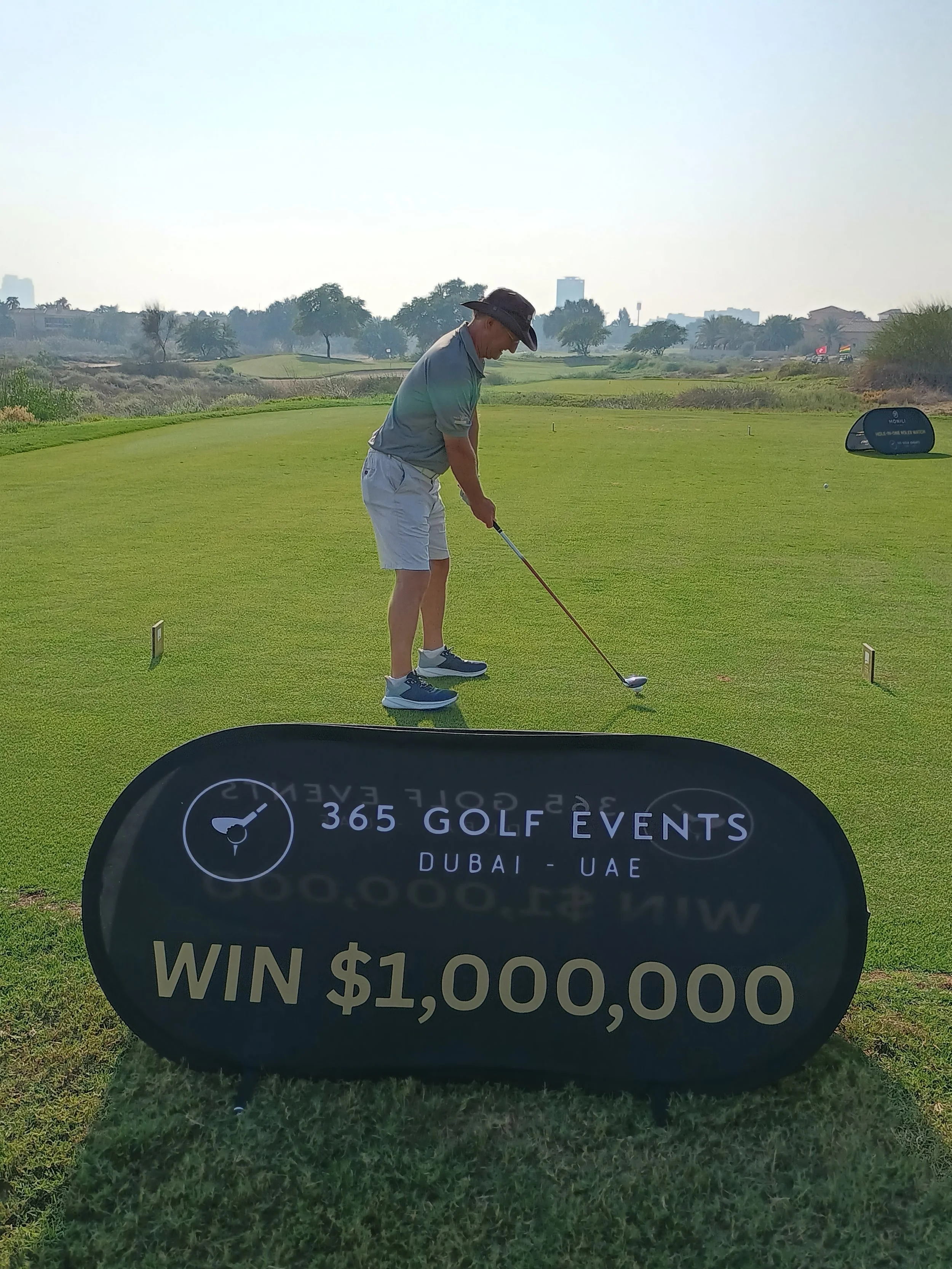 A man playing golf on a golf course in Dubai, UAE, with a sign advertising a golf event and a chance to win $1,000,000.