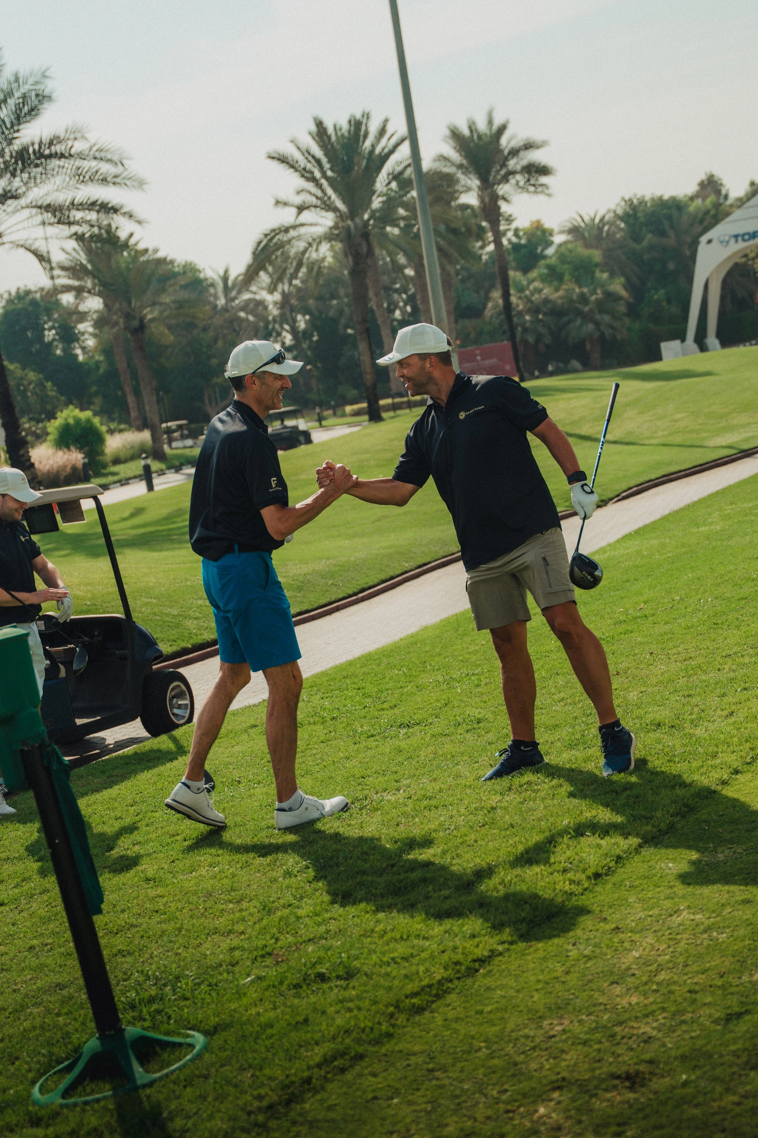 Two men are on a golf course, exchanging a handshake. One is holding a golf club, and a golf cart is nearby with another person sitting inside. The background has palm trees and a clear sky.