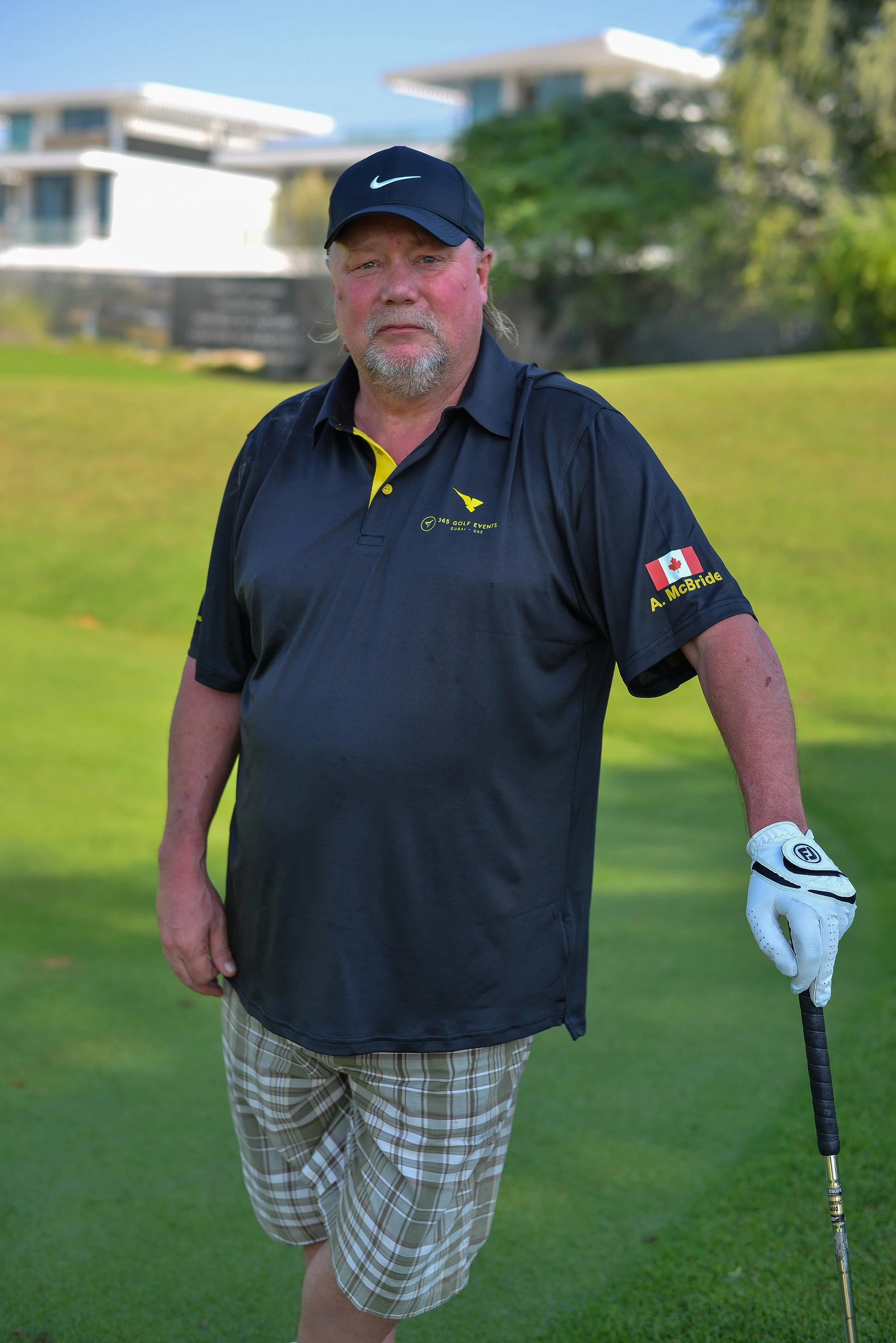 A middle-aged man standing on a golf course, wearing a black golf shirt, plaid shorts, and a black cap, holding a golf club with a golf glove on his right hand.