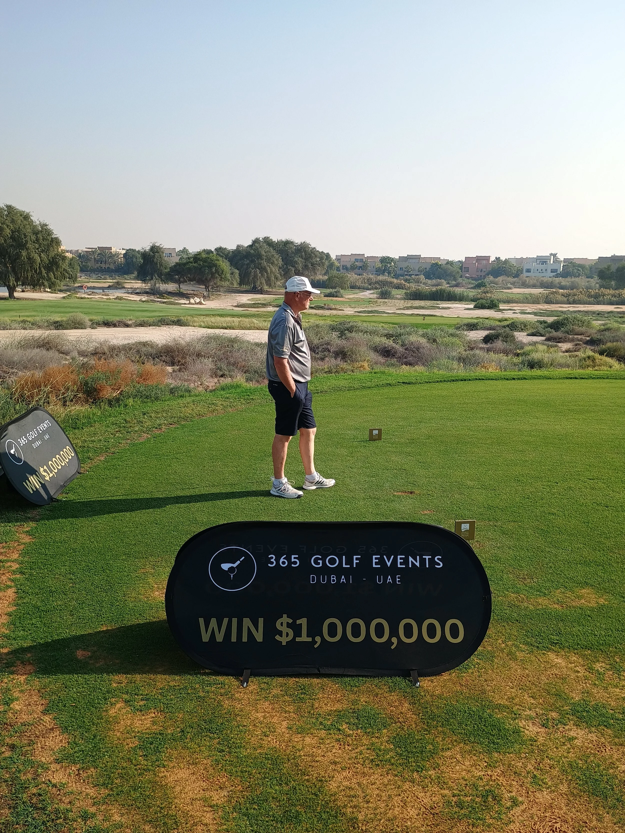 A man on a golf course near a golf tee, with signage advertising a golf event in Dubai, UAE, offering a $1,000,000 prize.