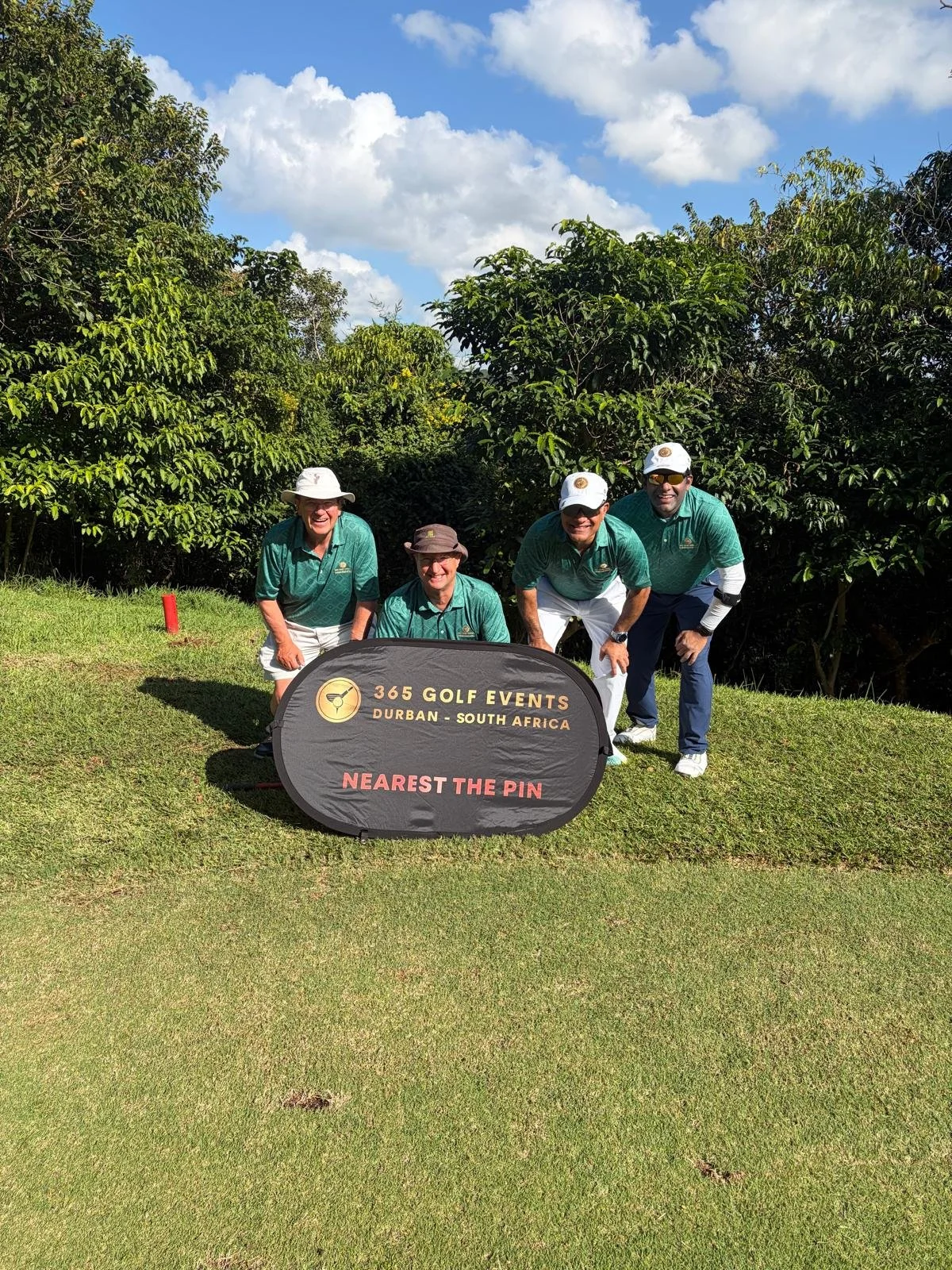 Four men in green shirts and hats kneel and stand behind a sign on a golf course with trees and a blue sky in the background.