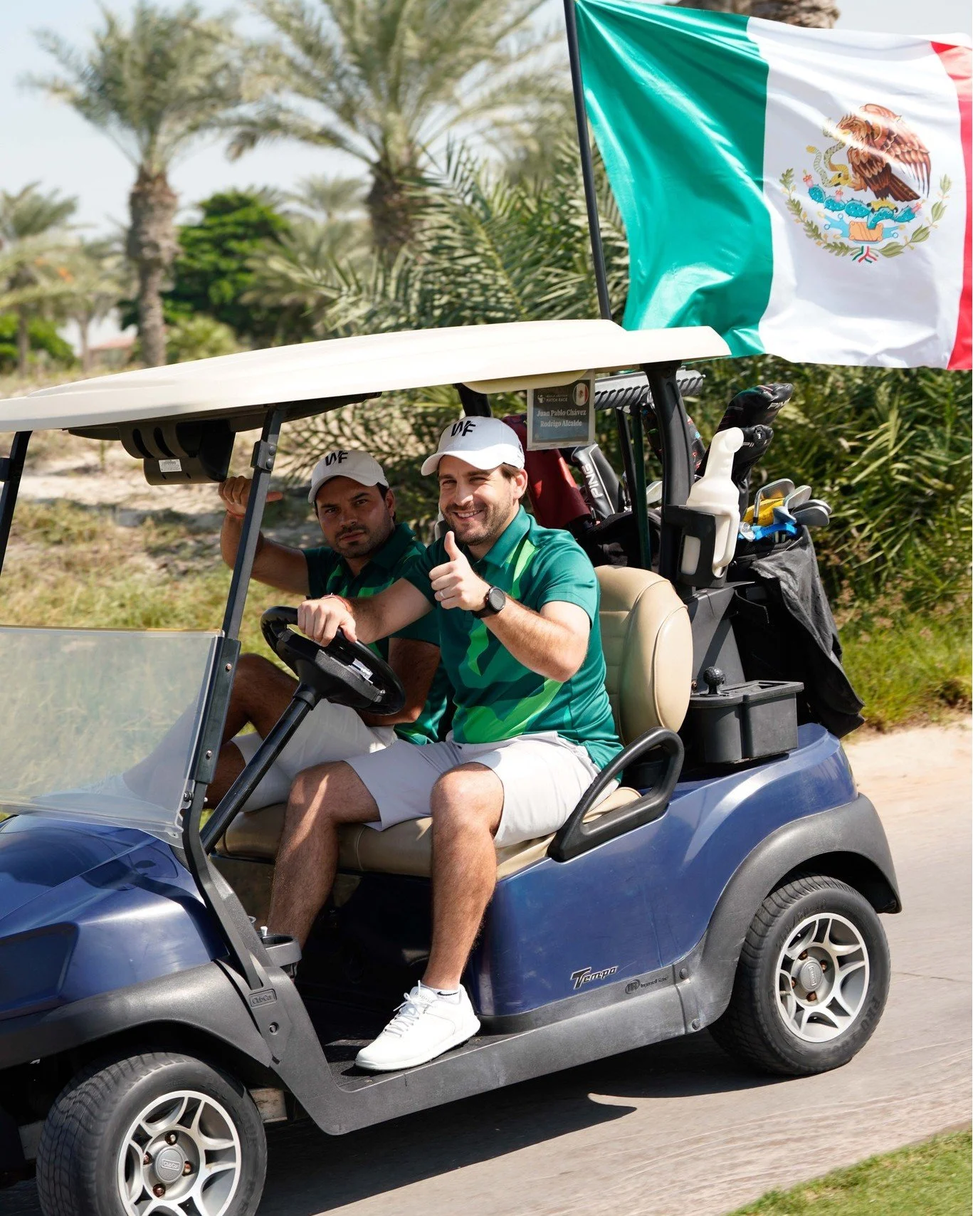 Two men riding in a blue golf cart with a Mexican flag attached, with green trees and palm trees in the background. One man is driving and giving a thumbs-up, both wearing green shirts and white caps.