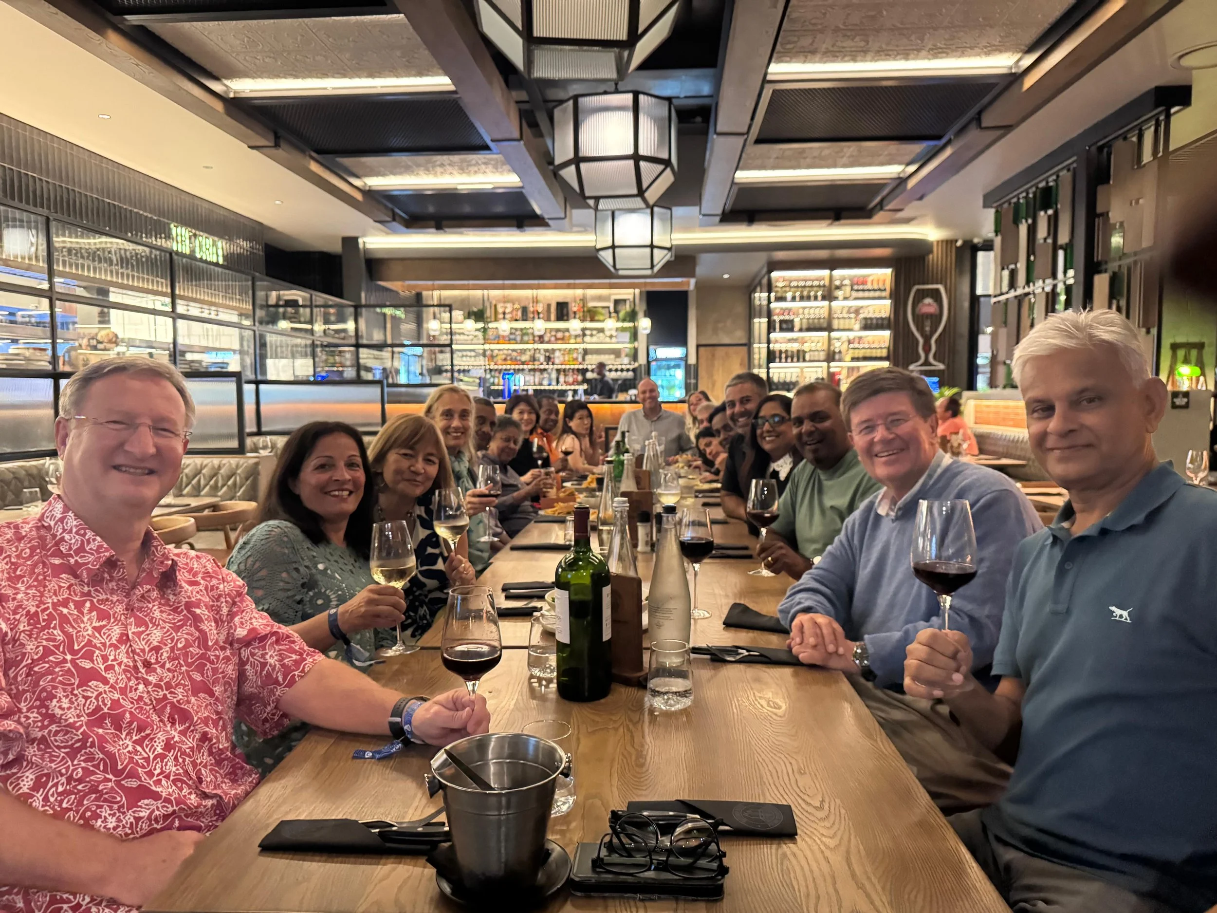 A diverse group of people sitting around a long table in a restaurant, smiling and holding glasses of wine, with a ceiling decorated with modern lighting fixtures and shelves stocked with bottles in the background.