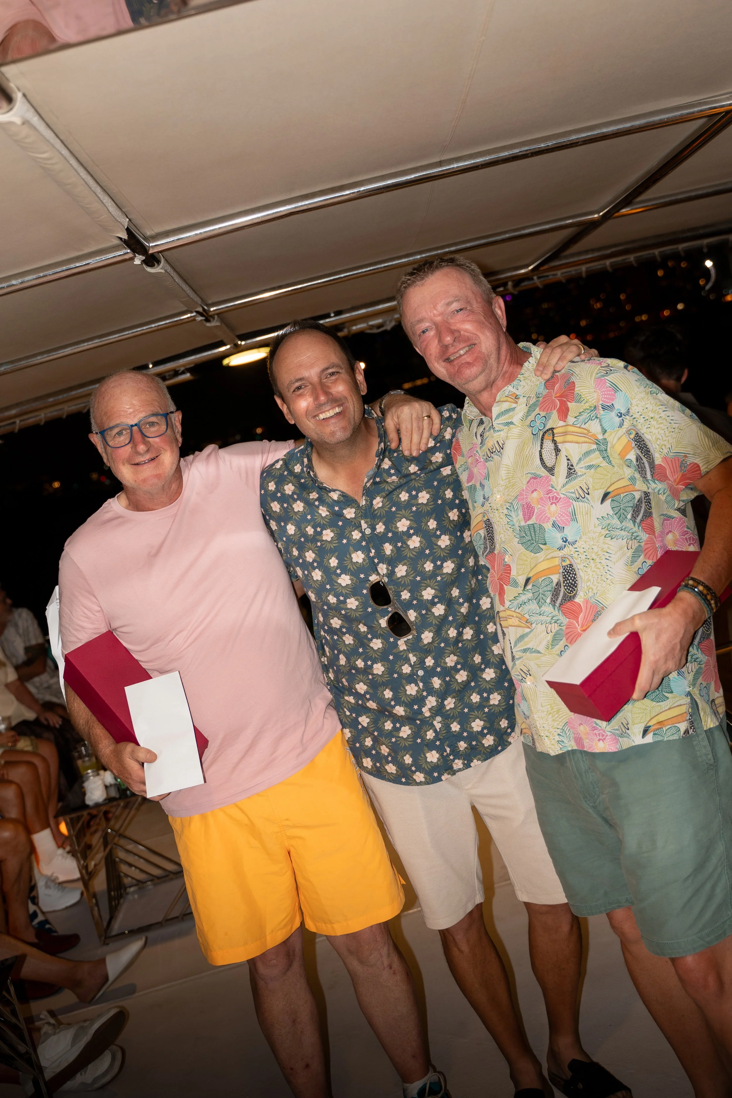 Three smiling men with light skin standing close together, wearing colorful casual summer shirts and shorts, holding wrapped gifts, at an outdoor gathering during the night.