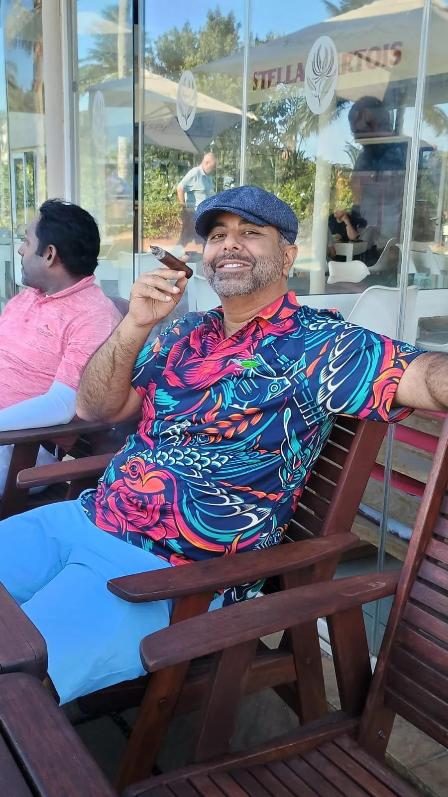 A middle-aged man wearing a blue cap and a colorful floral shirt sitting on a wooden chair at an outdoor cafe, holding a cigar, smiling at the camera.