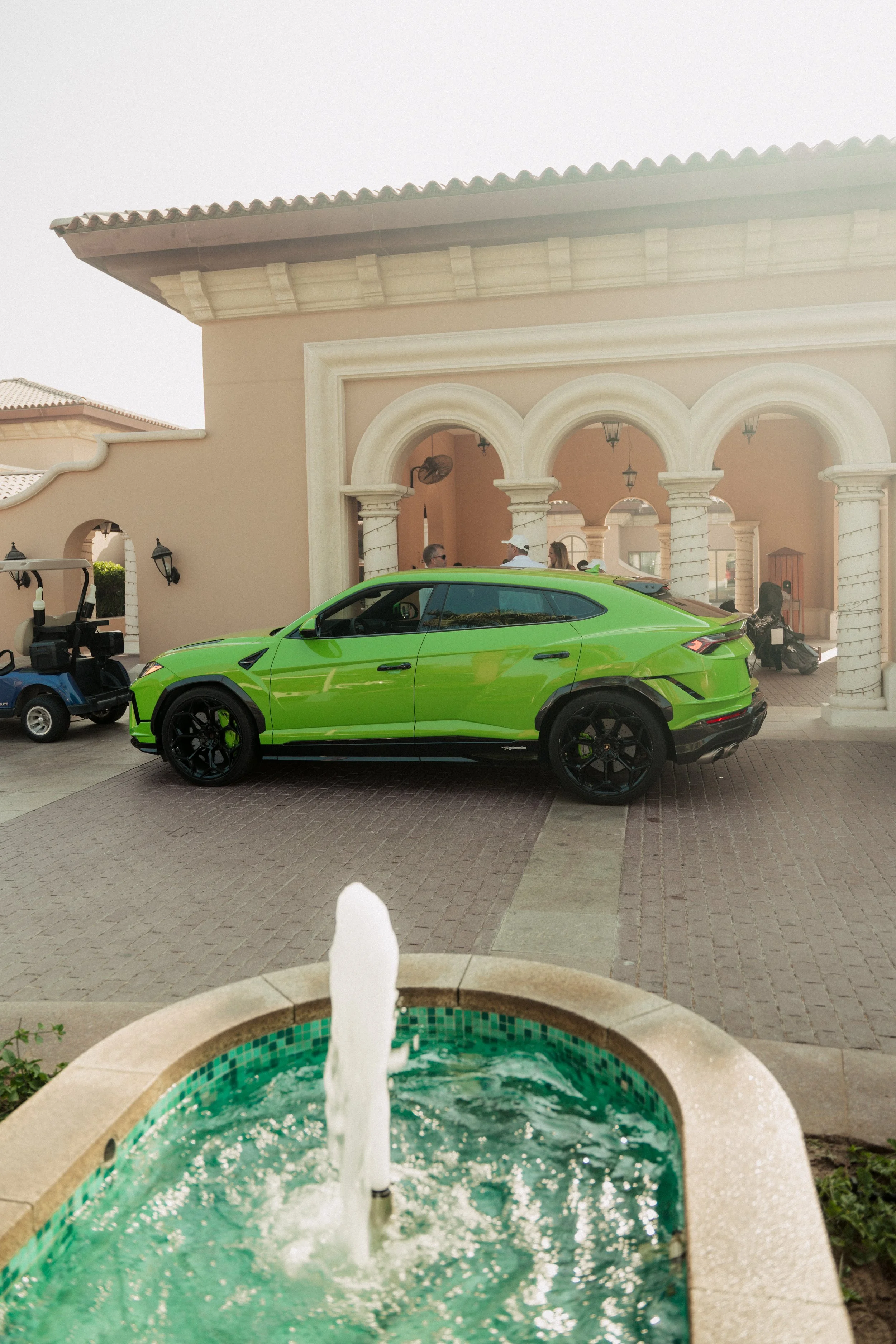 A bright green luxury SUV parked in front of a Mediterranean-style building with arches and columns. A small fountain with flowing water is in the foreground, and a golf cart is nearby.