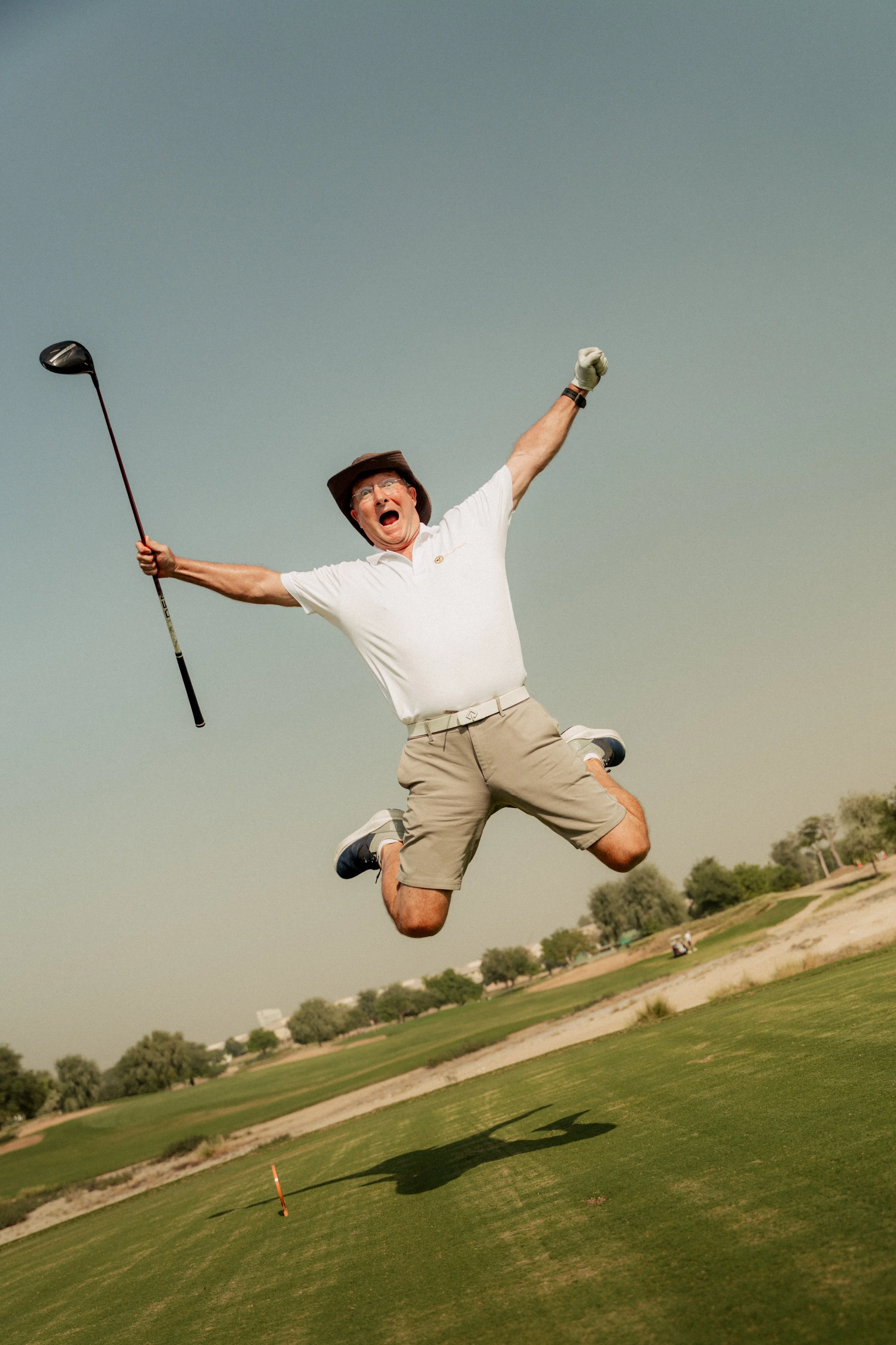 A man in golf attire jumping in the air on a golf course, holding a golf club in one hand, with a surprised or excited expression.