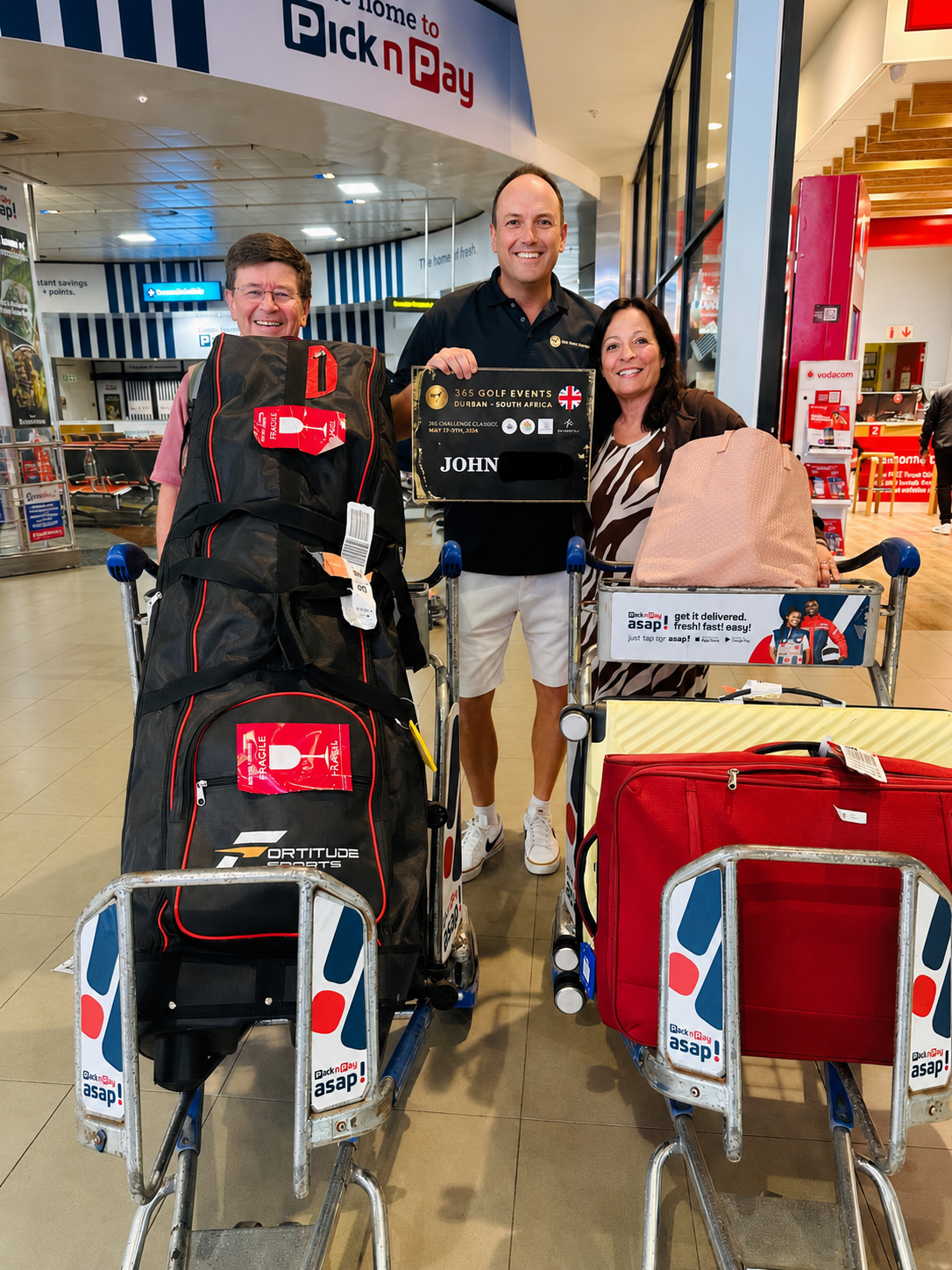 Three people standing with luggage carts at an airport, smiling. One man holds a sign for a golf event in South Africa, with bags on the carts including a large black golf bag, a red suitcase, and a pink bag.