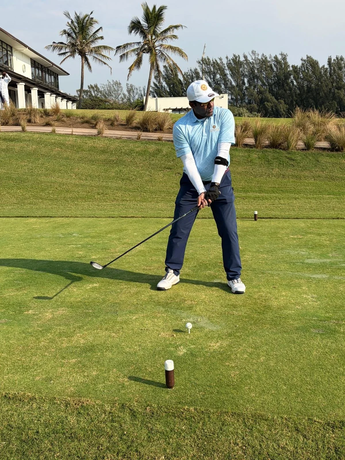 A man playing golf on a green course, preparing to hit the golf ball with a club, dressed in a light blue shirt, dark blue pants, and a white cap, with palm trees and a building in the background.