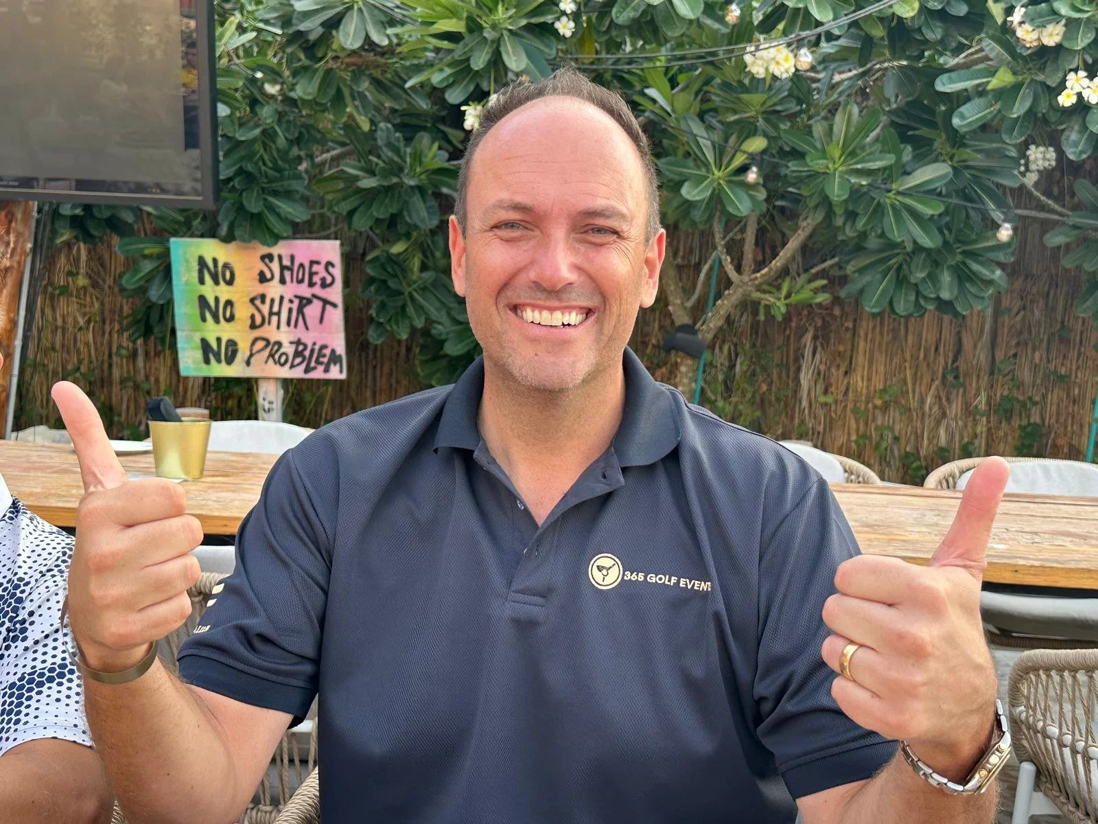 A smiling man in a navy polo shirt giving two thumbs up, seated outdoors with lush greenery and a colorful sign in the background that reads 'No shoes, no shirt, no problem'.