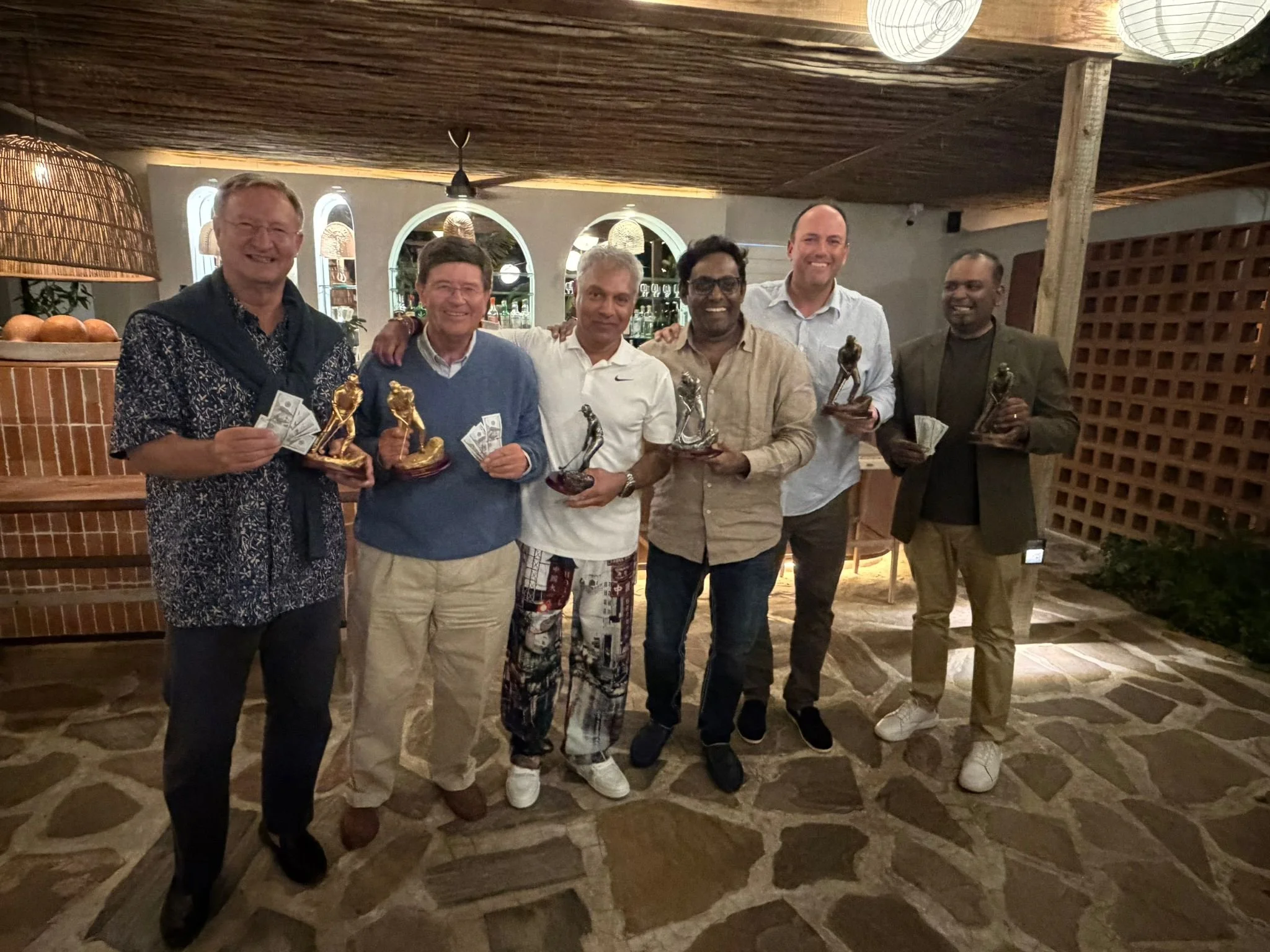A group of six men standing indoors in front of a bar, holding trophies and cash. They are smiling and dressed in casual and semi-formal attire.