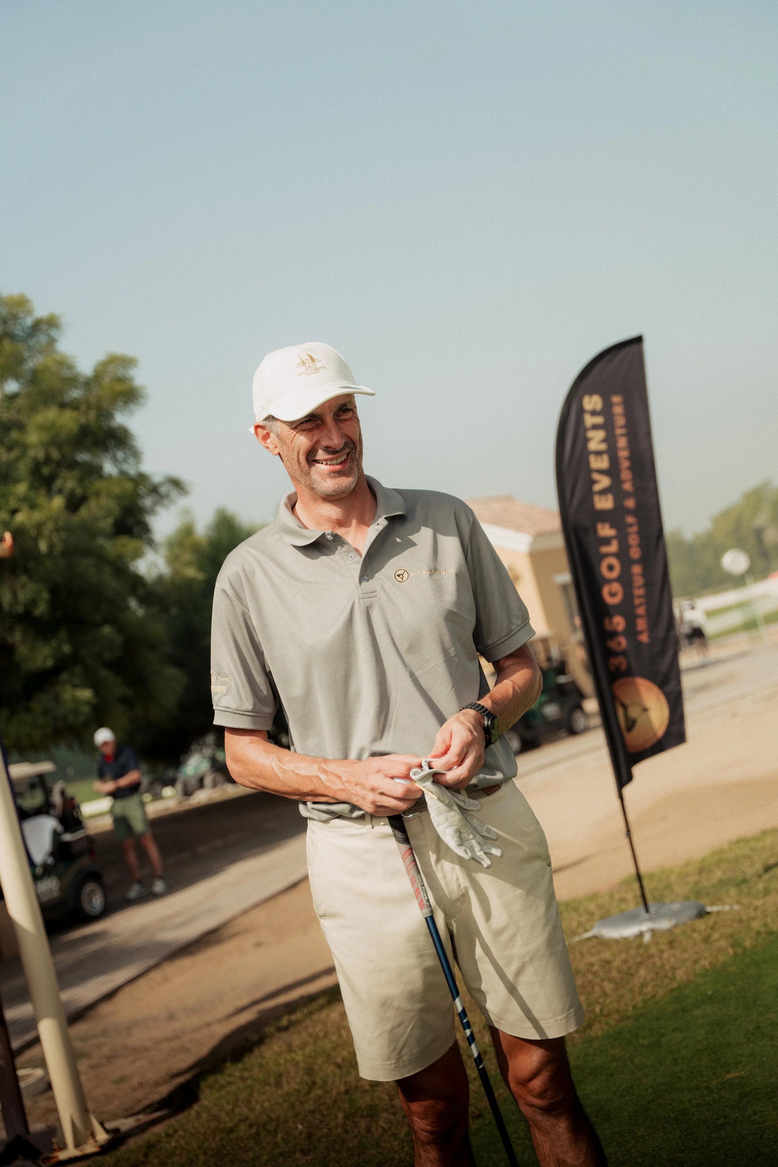 A man smiling and holding a golf club on a golf course during the day, wearing a white cap, gray polo shirt, and khaki shorts. A black promotional flag with orange text is behind him, and another person is visible in the background near golf carts.