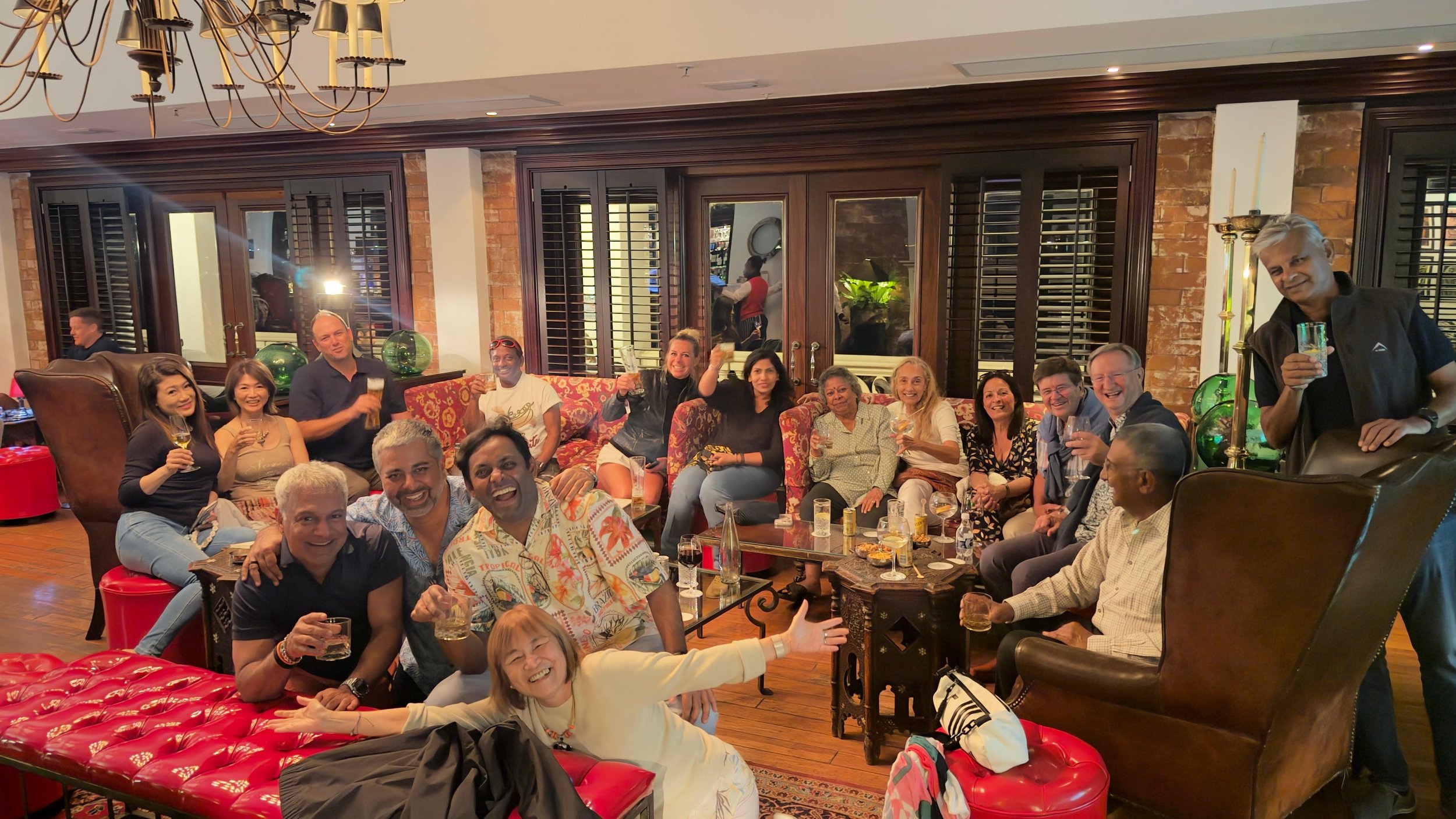 A group of diverse people gathering in a cozy, warmly lit living room, smiling and holding drinks, some sitting on red and floral-patterned furniture and others standing, with large windows and wooden accents in the background.