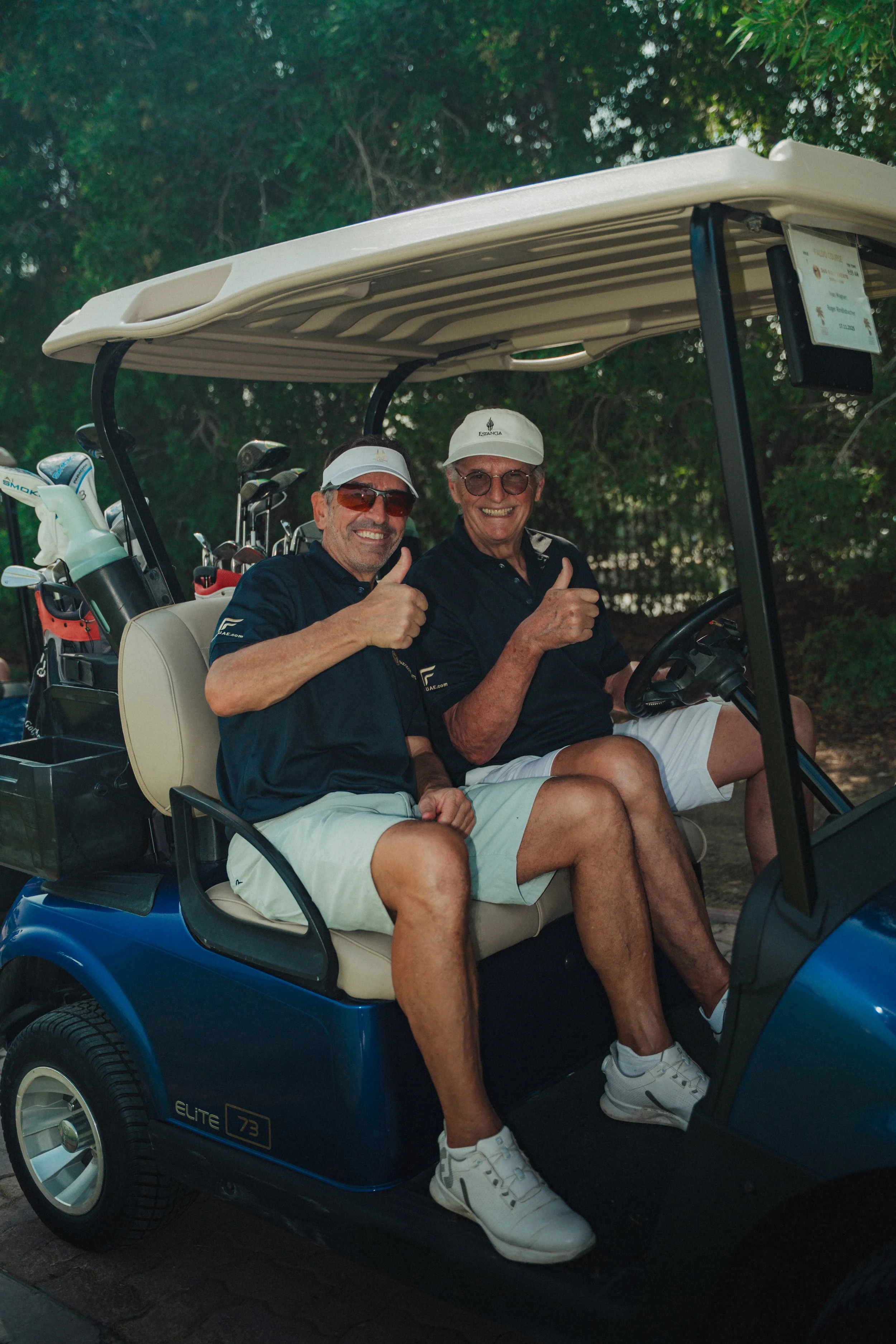 Two men sitting in a golf cart, smiling and giving thumbs up. They are dressed in golf attire with hats and sunglasses, and there are golf clubs in the back of the cart.