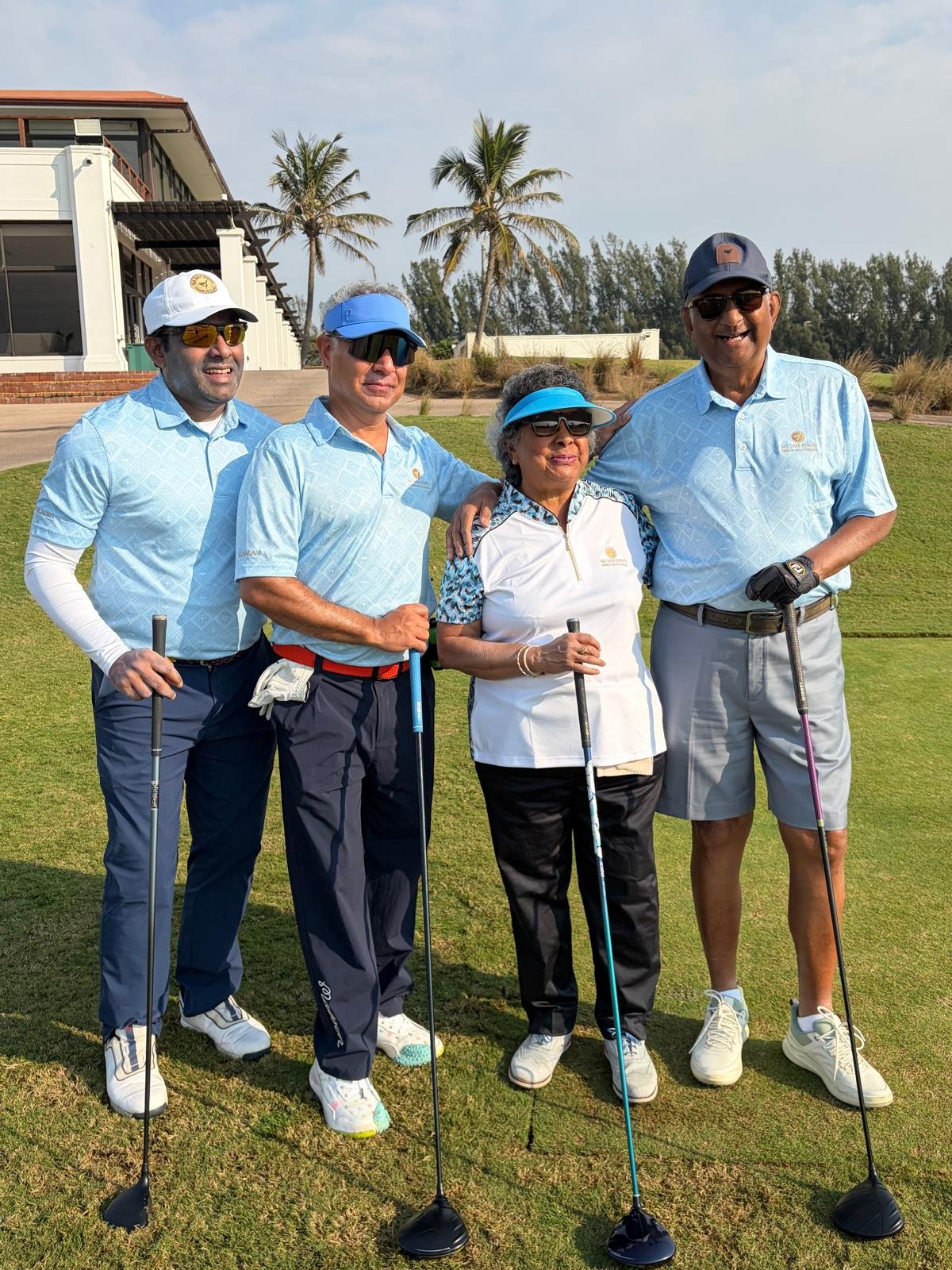 Four adults standing on a golf course with golf clubs, smiling, and wearing golf attire with an ocean view and palm trees in the background.