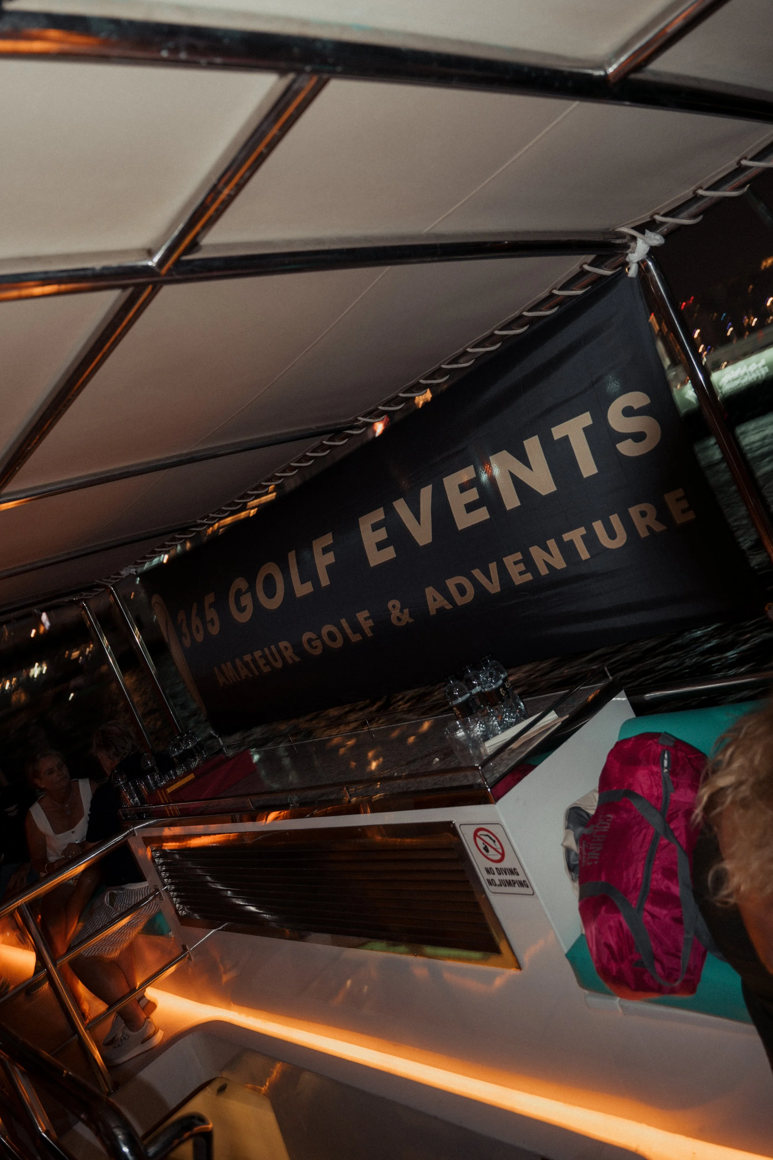 Interior of a boat with a banner displaying '365 Golf Events Amateur Golf & Adventure' and a woman sitting at a table with a pink and gray backpack.