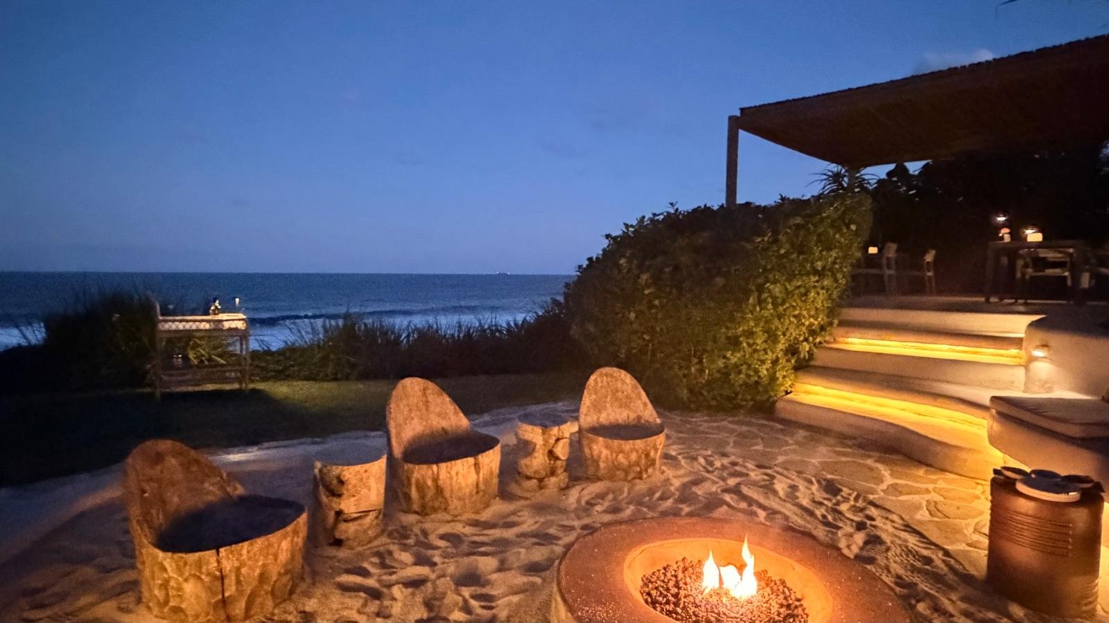 Beachside patio at night with a firepit, wooden chairs, stairs with lighting, overlooking the ocean, with a bench and bushes.