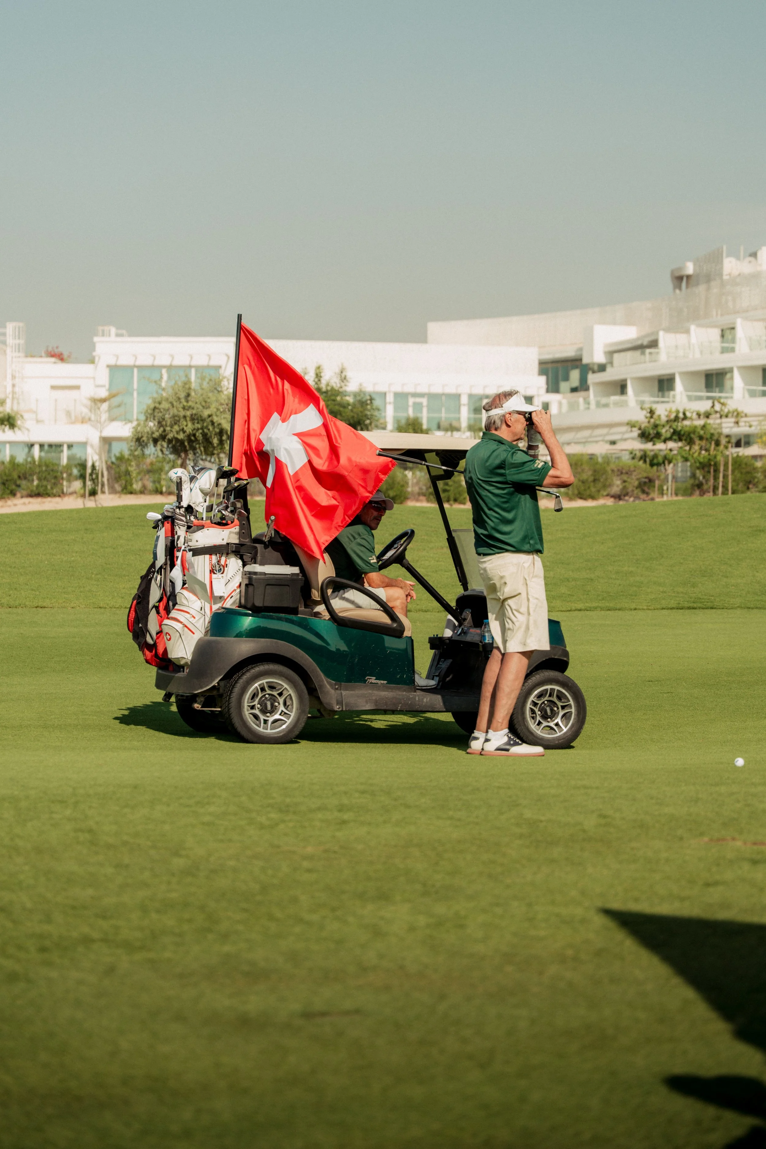 A golfer standing next to a golf cart with a Swiss flag, on a golf course, adjusting his visor while a person seated in the cart observes.