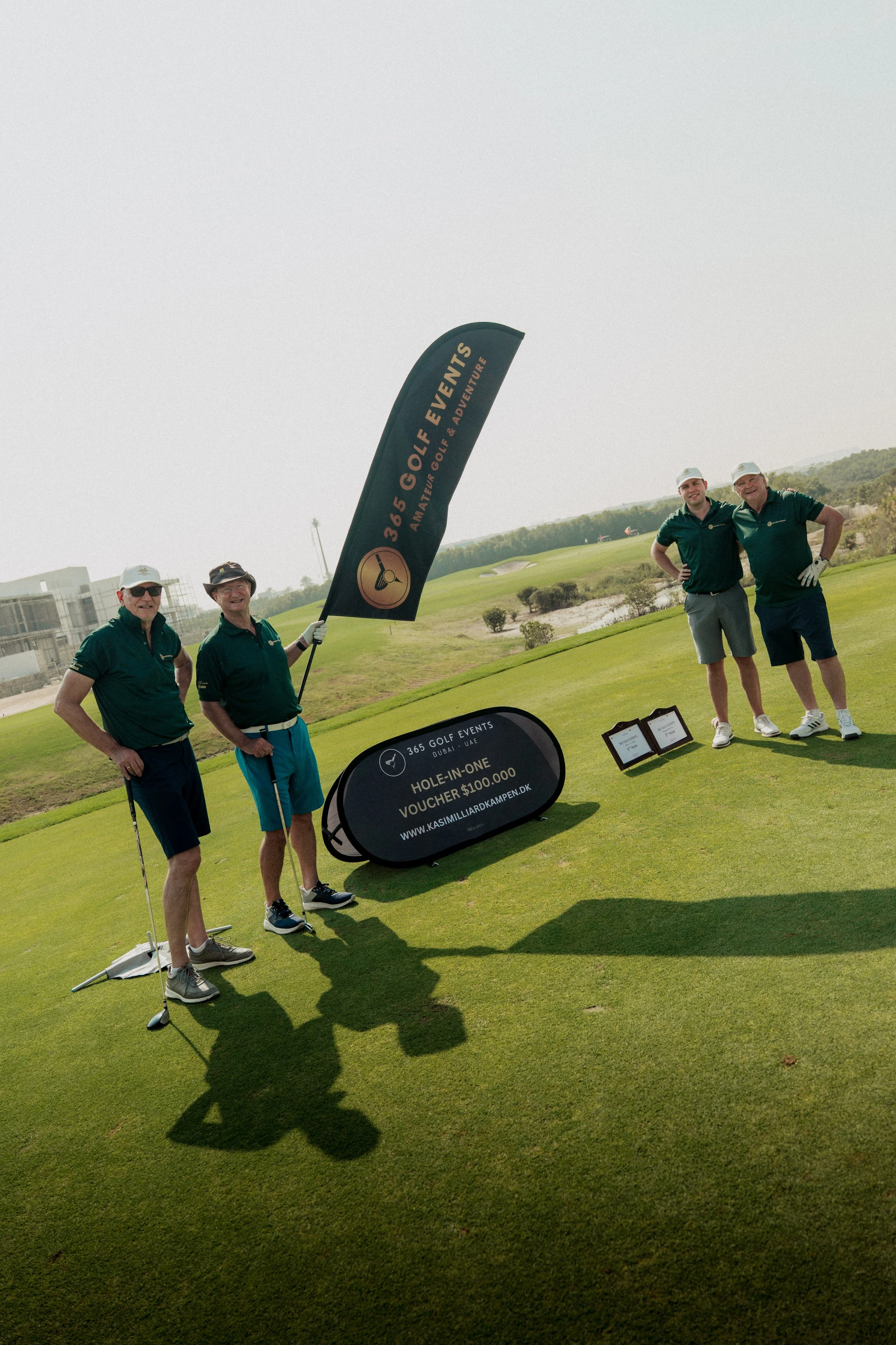 Four men on a golf course celebrating a hole-in-one achievement, with a large sign promoting a golf event and a voucher prize.