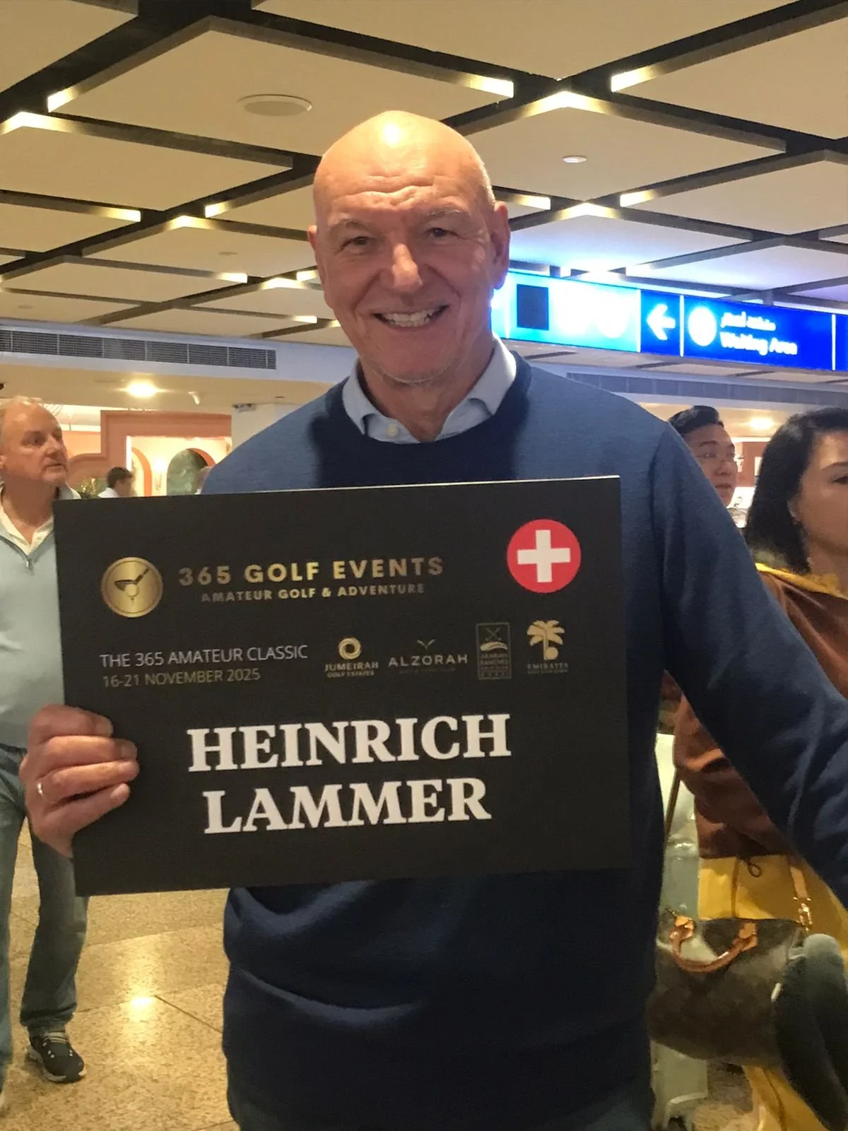 Man smiling at an indoor event holding a sign with event details, with other people in the background.