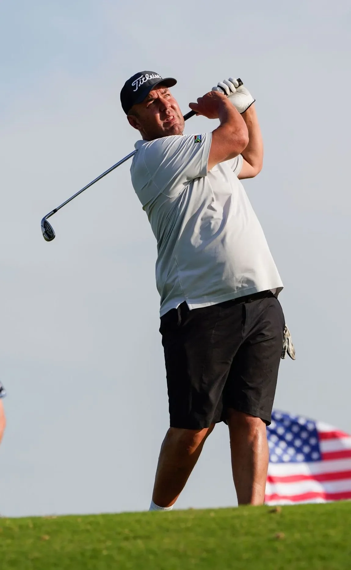 Man playing golf, swinging golf club on a green field with American flag in the background.