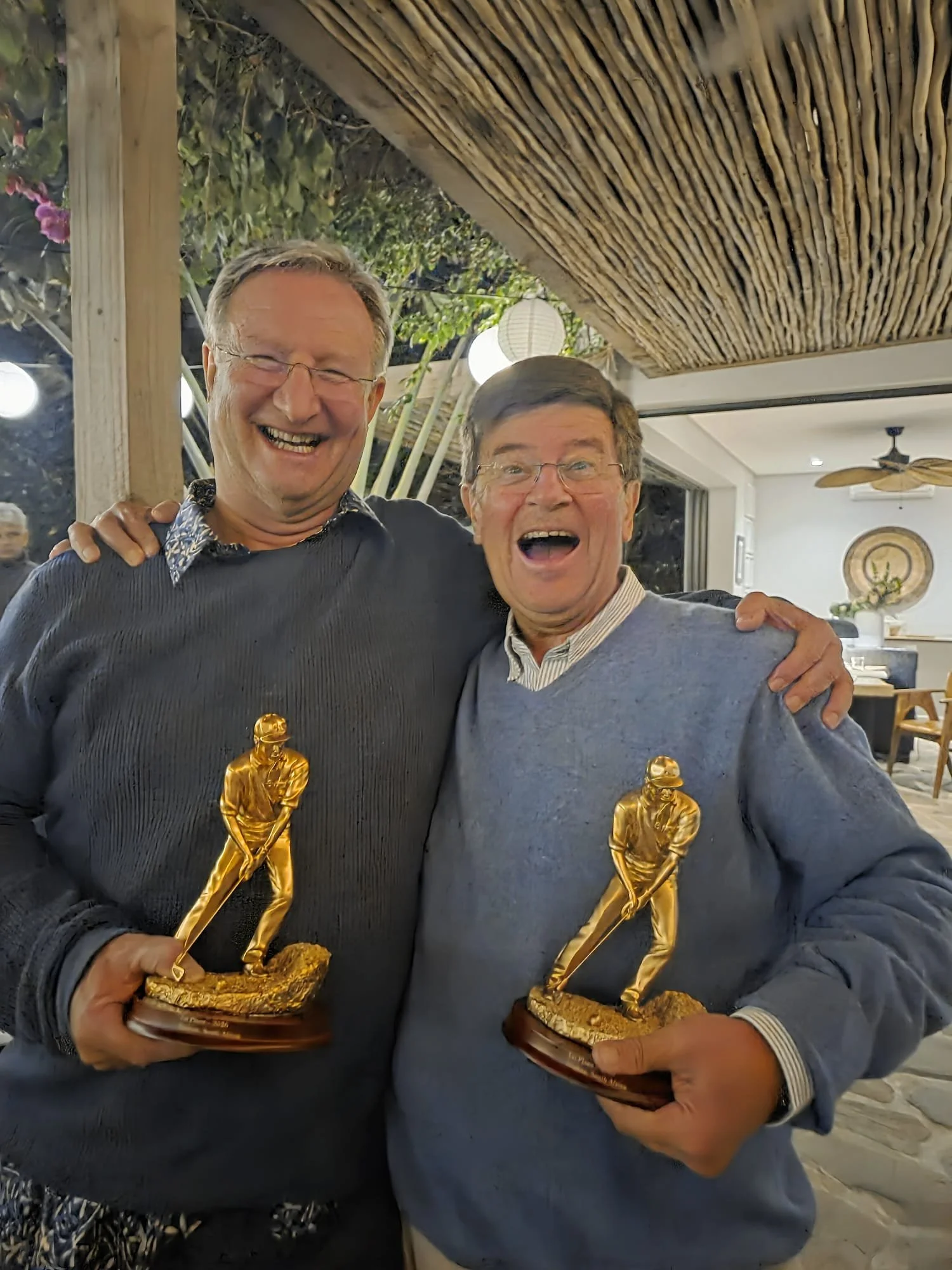 Two men smiling and holding Goldman Award trophies indoors, with a ceiling fan and decorative wall plate in the background.