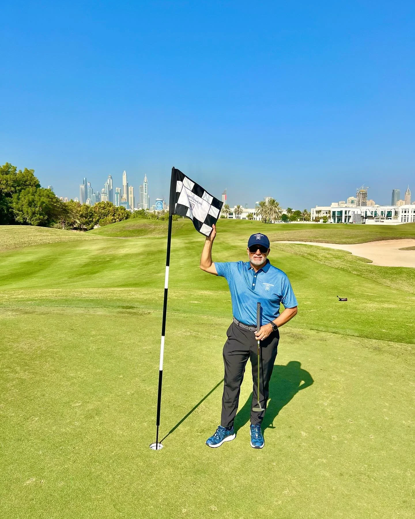 Man in blue shirt and black pants holding a checkered golf flag on a golf course with city skyline in the background.