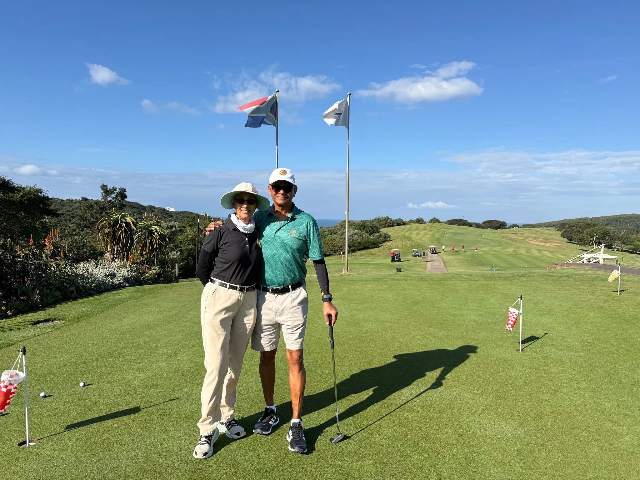 A man and woman standing together on a golf course, smiling, in front of a putting green with flags, with golf clubs and balls around them, and trees and hills in the background under a blue sky with some clouds.