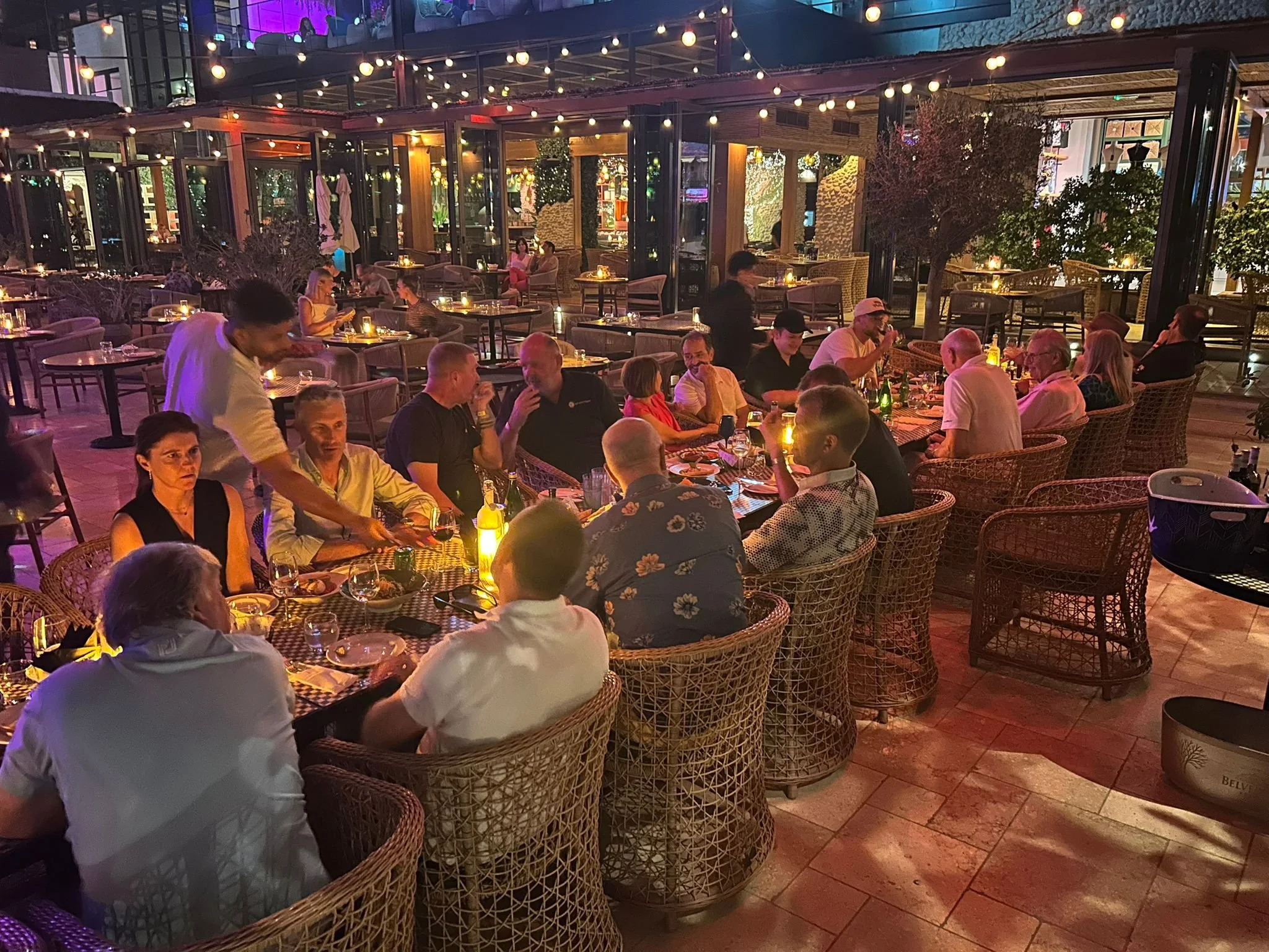 People dining inside a restaurant with string lights and outdoor view, with tables filled with food, drinks, and candles.