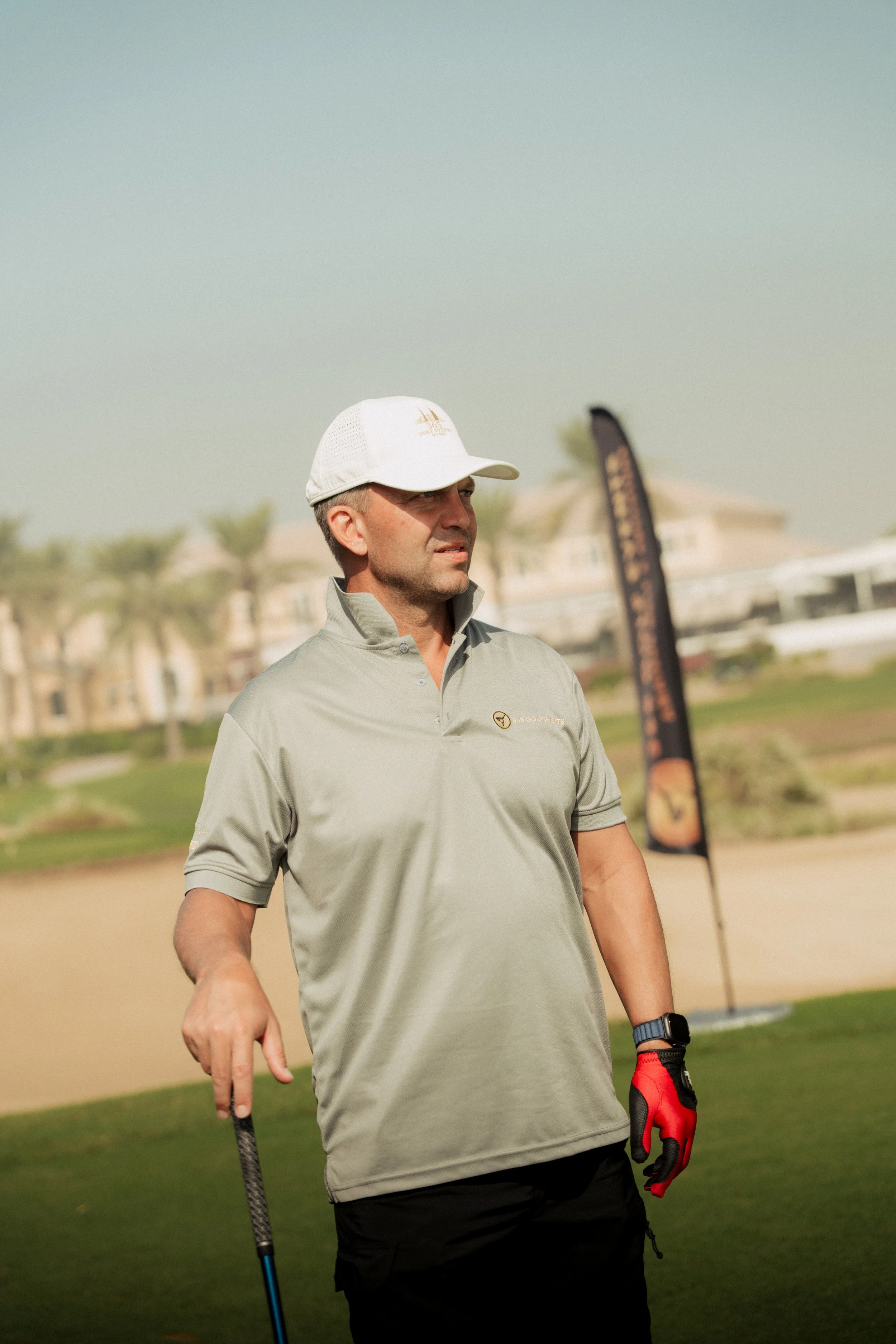 A man in golf attire standing on a golf course holding a golf club, wearing a white cap and a gray polo shirt, with a flag and residential buildings in the background.