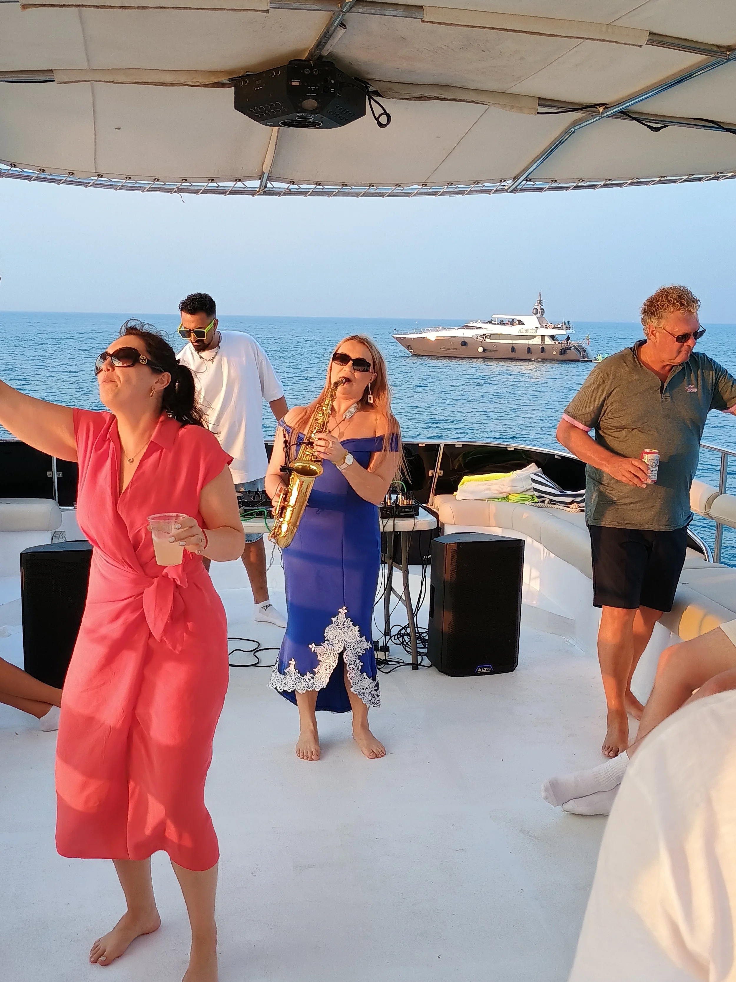 Group of people enjoying a boat party with a woman playing the saxophone, dancing, and relaxing on a yacht on the water during sunset.