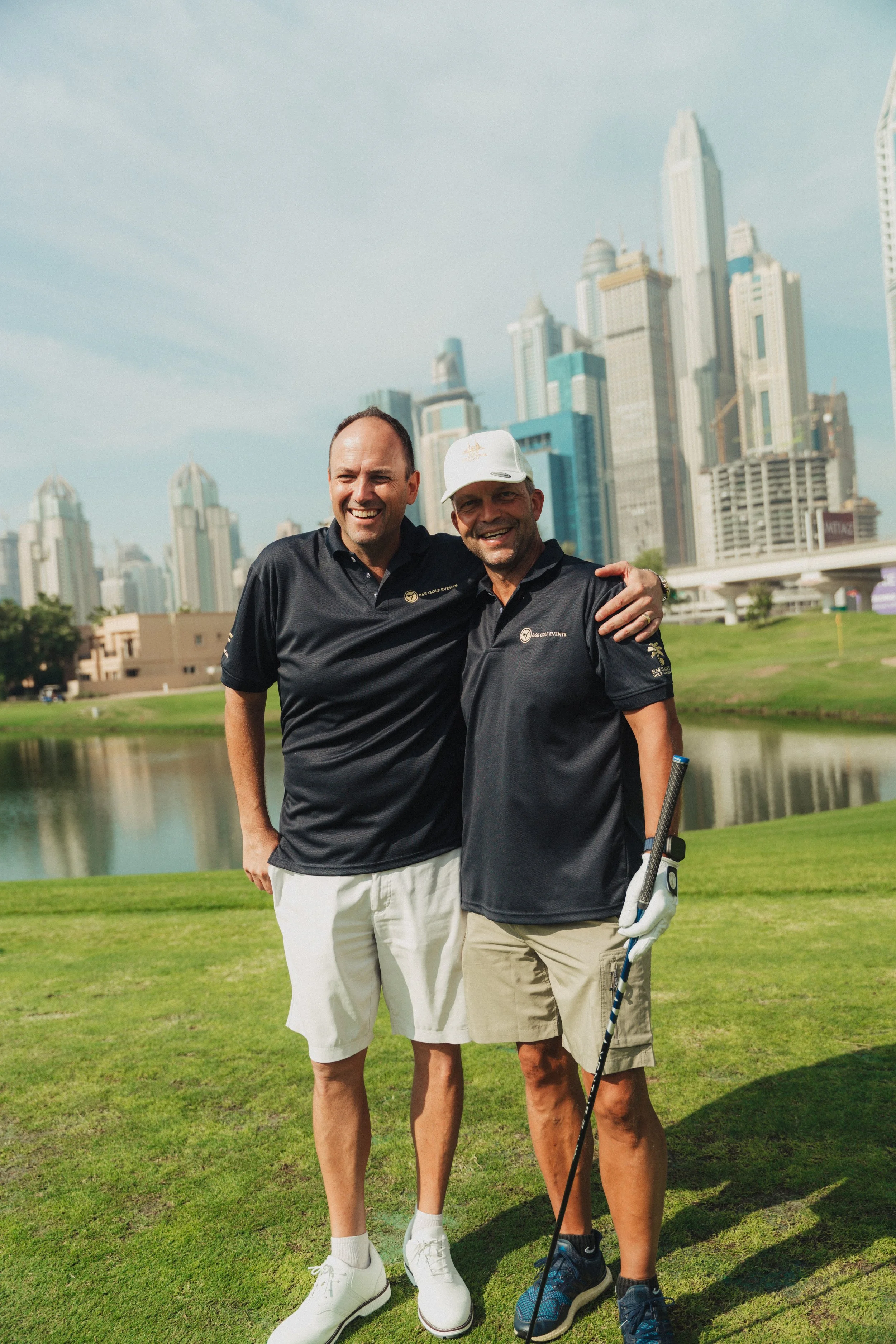 Two men standing on a golf course with a city skyline in the background, smiling and posing for a photo.