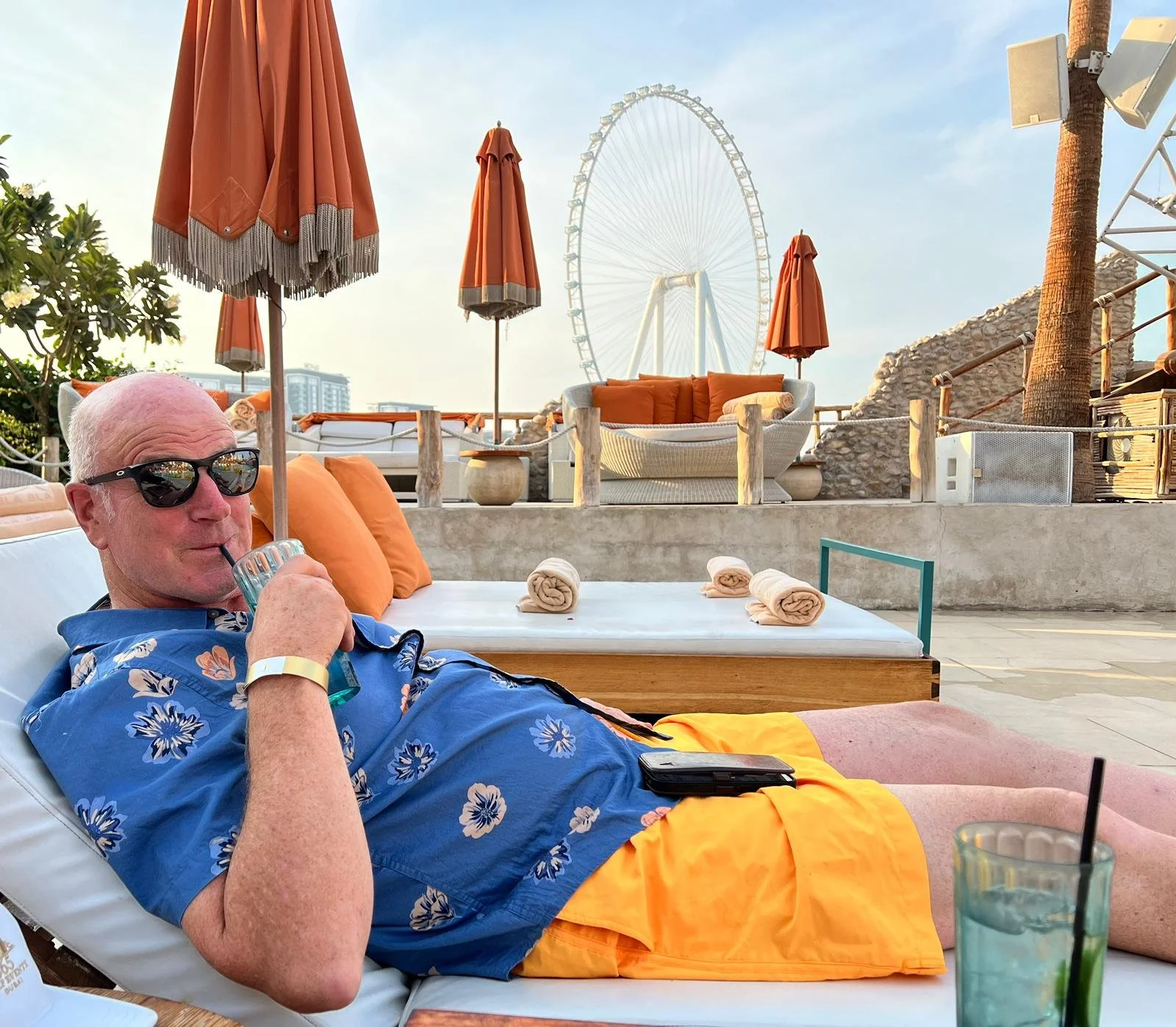 A man in sunglasses, a blue Hawaiian shirt, and orange shorts relaxing on a lounge chair drinking a beverage with a straw, with a cityscape, Ferris wheel, umbrellas, and outdoor furniture in the background.