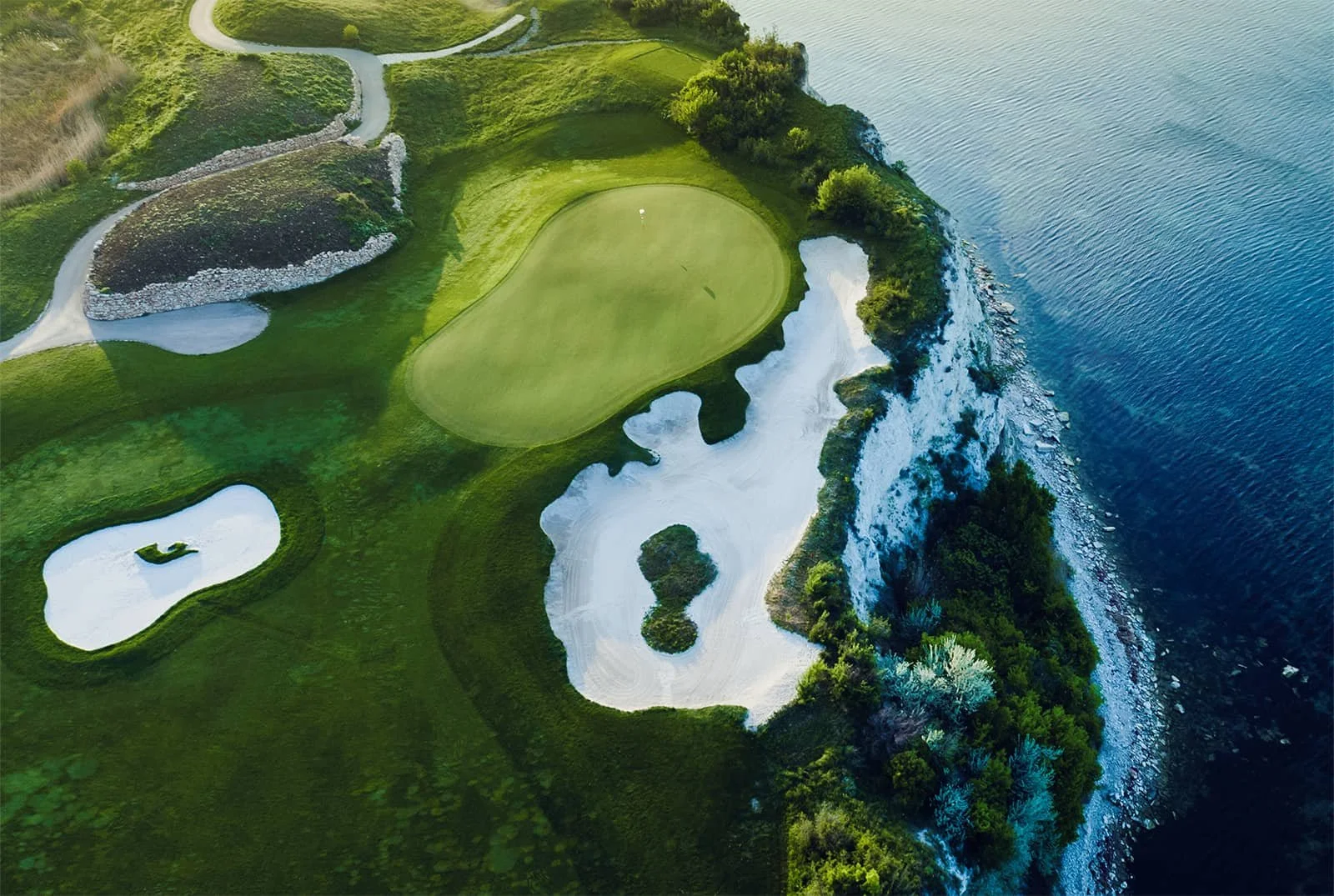 Aerial view of a golf course situated on a cliff overlooking a body of water, with lush green fairways, sand traps, and a putting green.